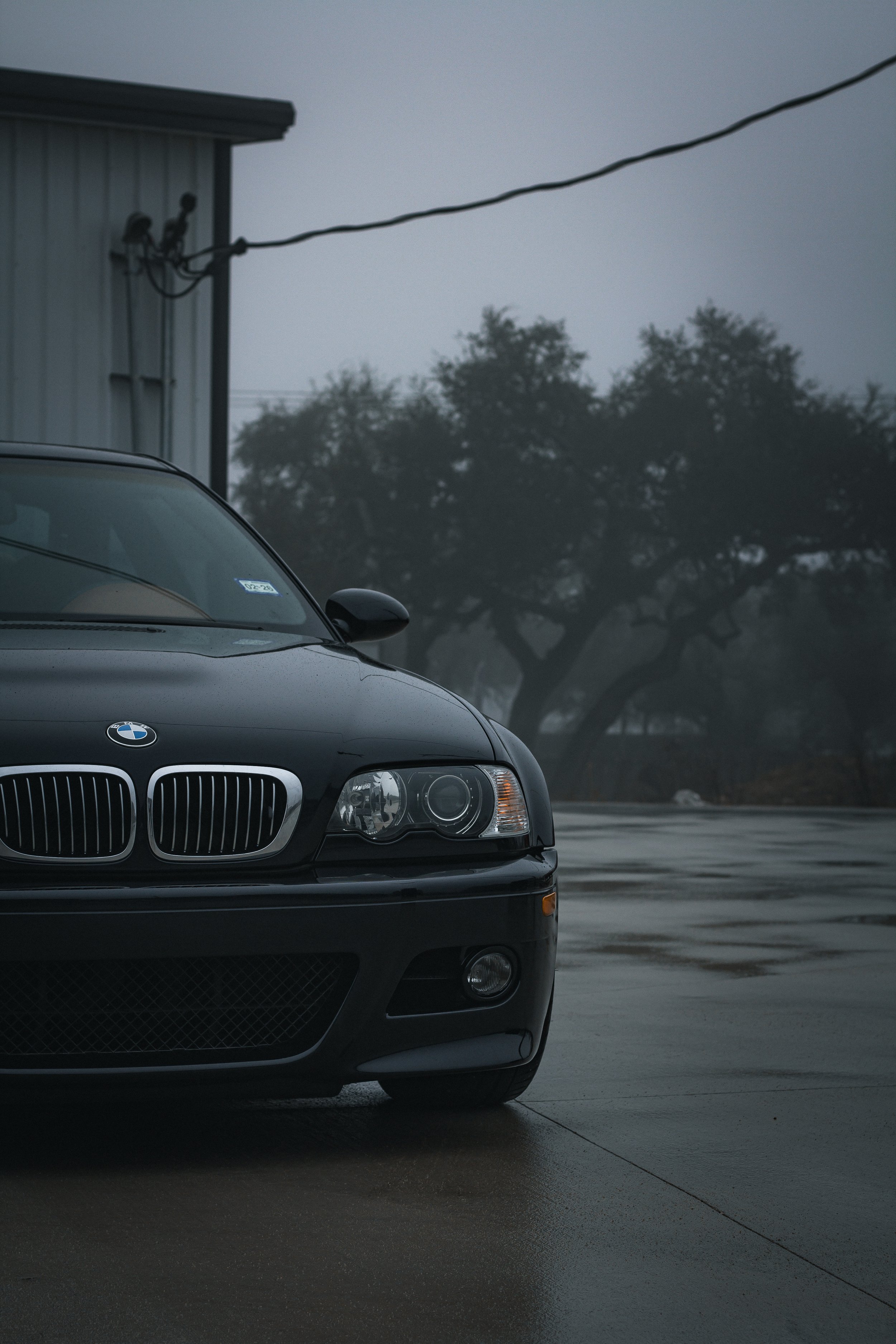Coche BMW negro estacionado en un condominio en un día lluvioso con cielo nublado y árboles en el fondo.
