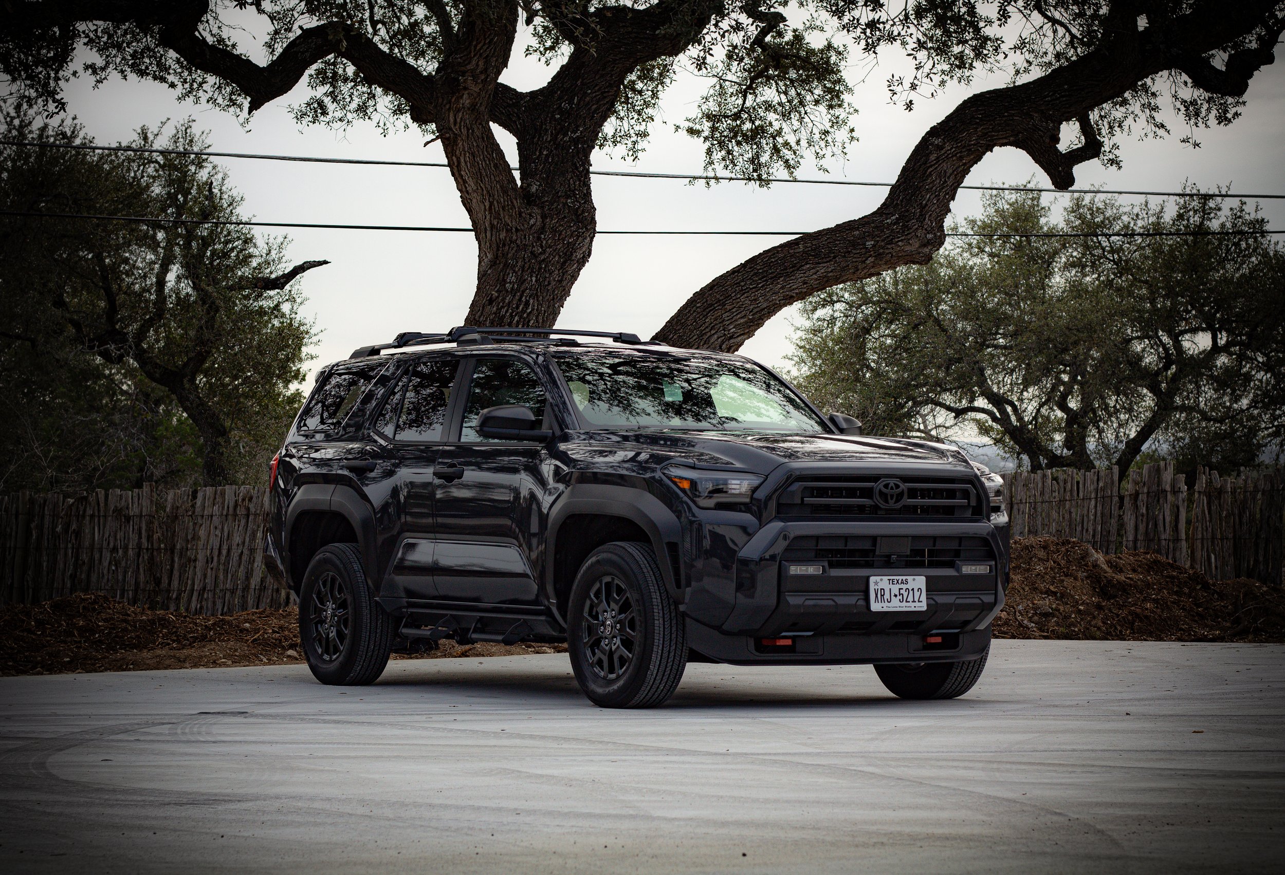 Un coche negro tipo SUV estacionado en un área abierta con árboles y un cercado de madera en el fondo.