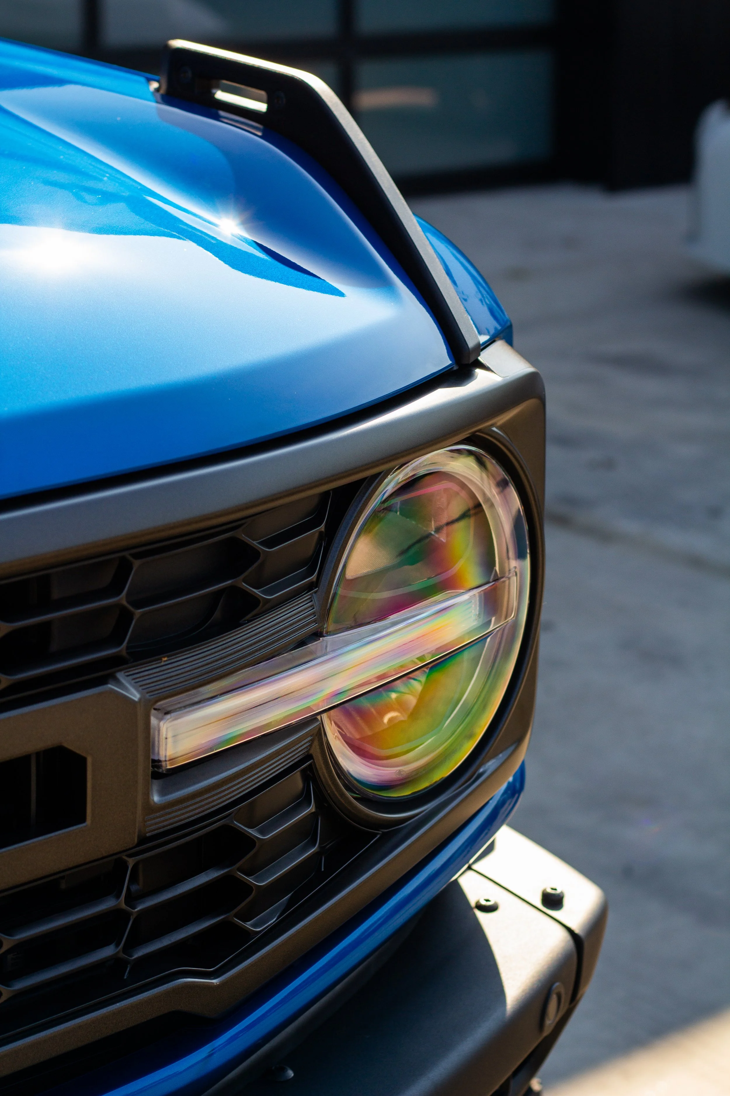 Primer plano del parachoques y faro de un coche azul, con el reflector del faro reflejando colores del arco iris.