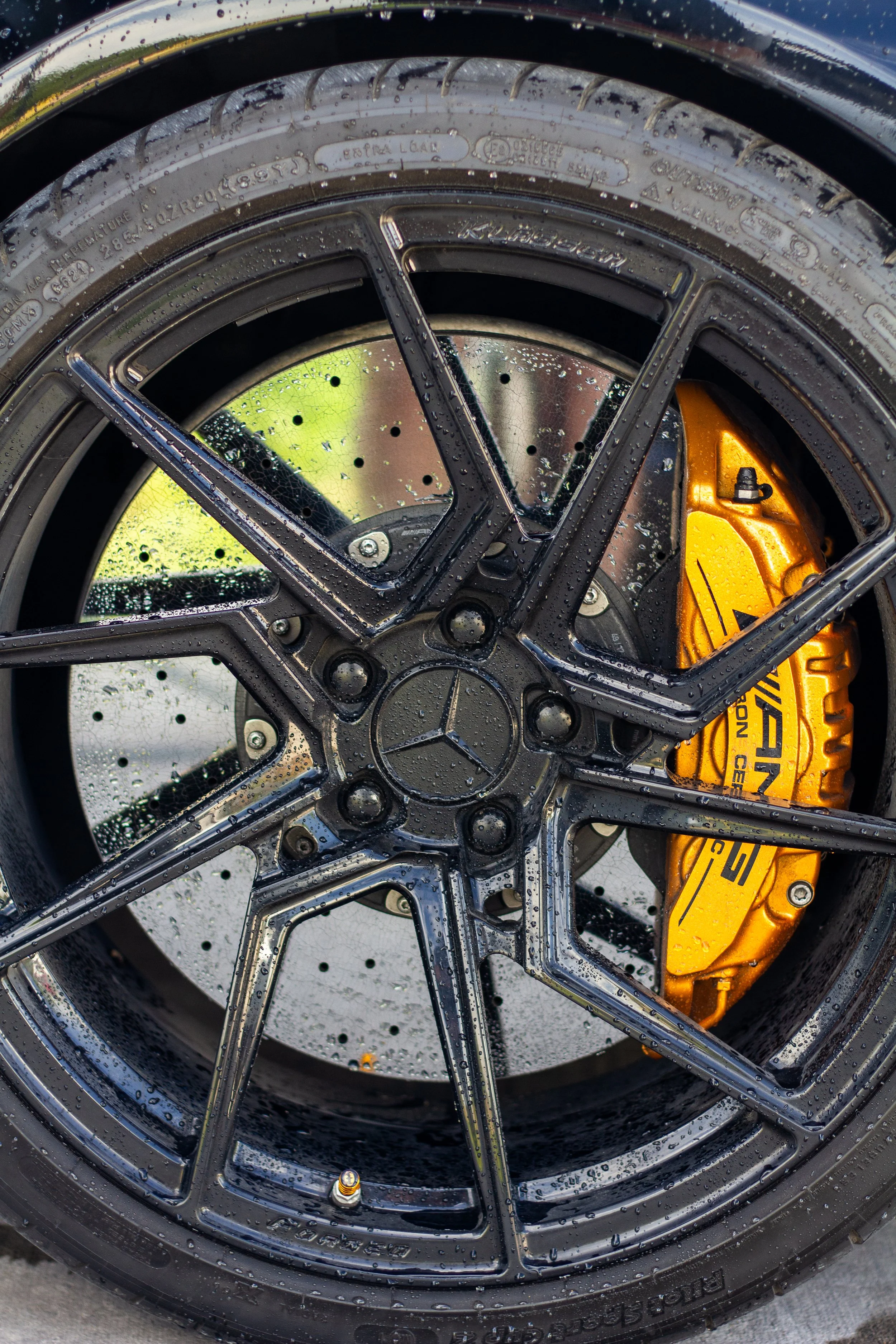 Rueda de un automóvil, con freno de disco dorado y llanta negra húmeda con gotas de agua.