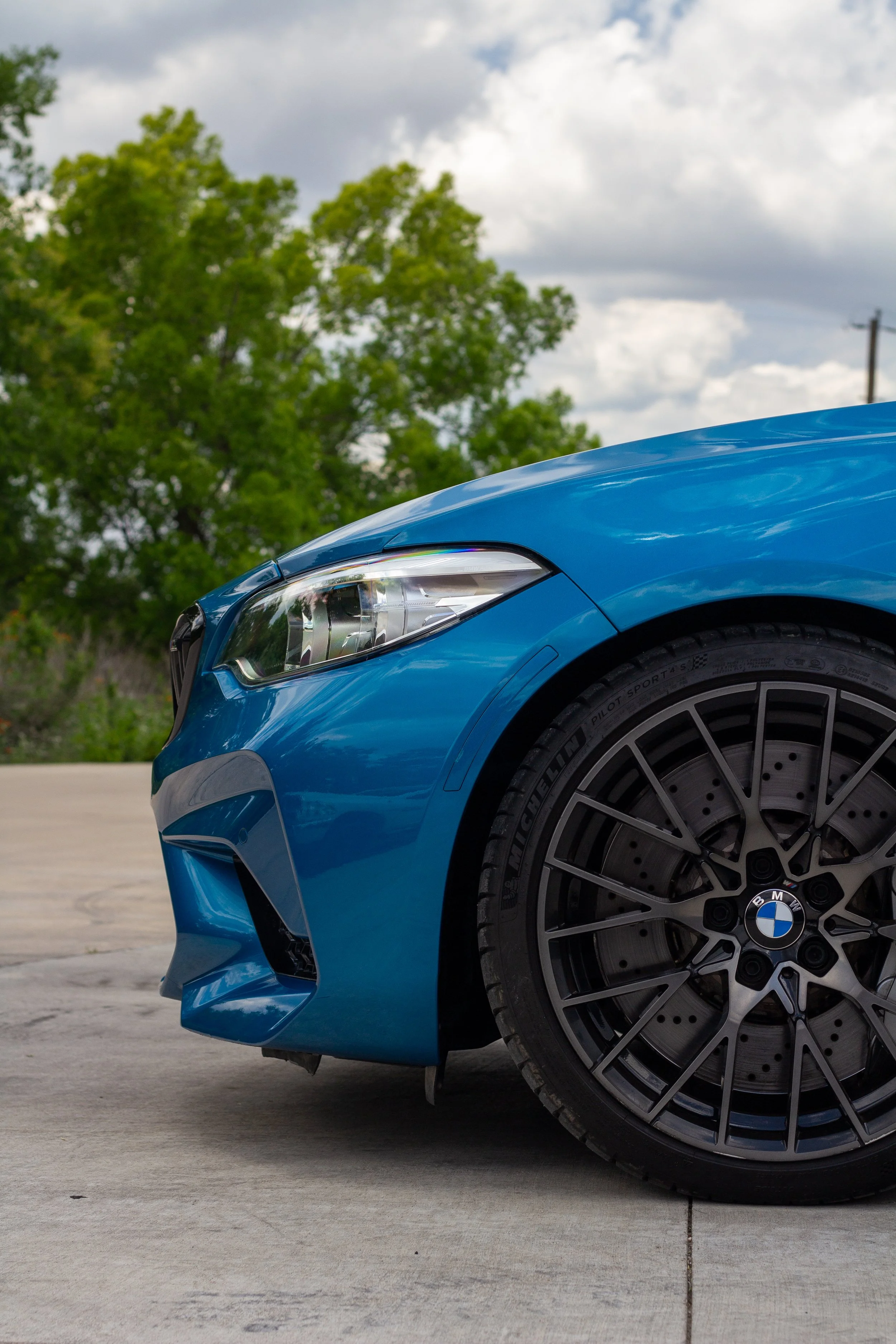 Frontal de un coche deportivo azul marca BMW en un lugar al aire libre con árboles y cielo nublado al fondo.