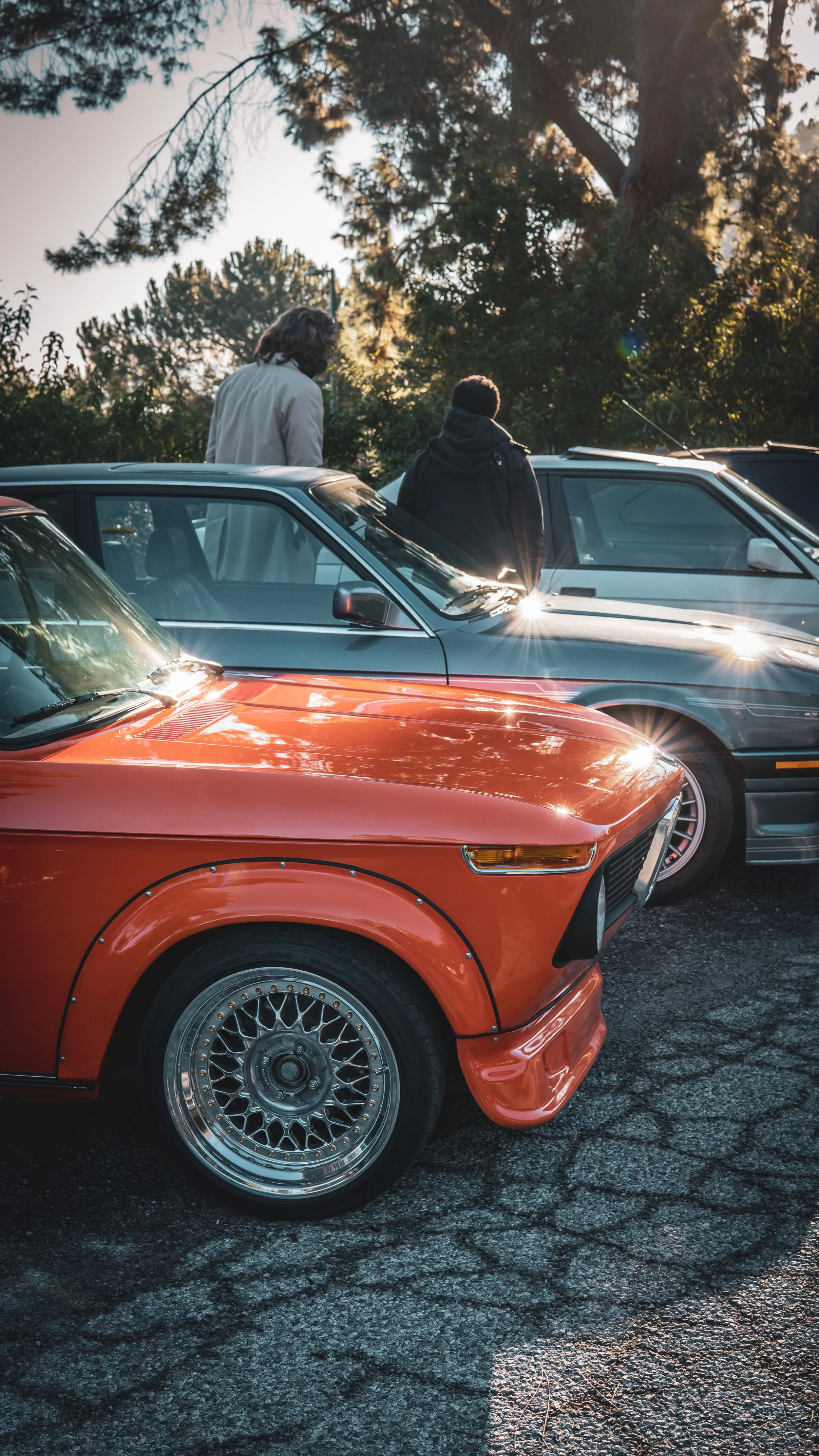 Dos autos deportivos antiguos, uno naranja y uno gris, estacionados en un parque, con dos personas de espaldas observando los autos durante el atardecer.