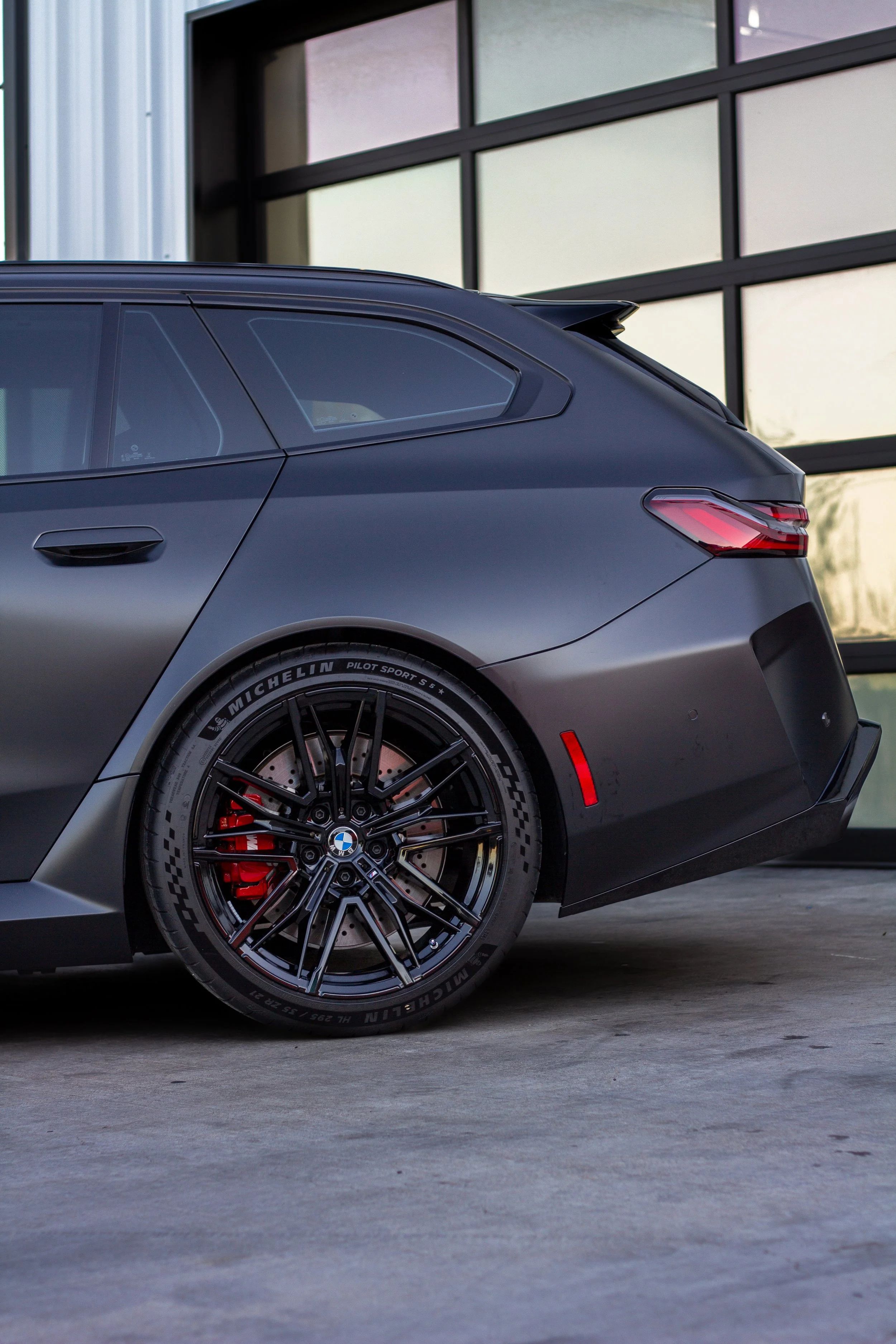 Coche deportivo BMW negro con neumáticos Michelin, en un estacionamiento junto a un edificio moderno con ventanas grandes.