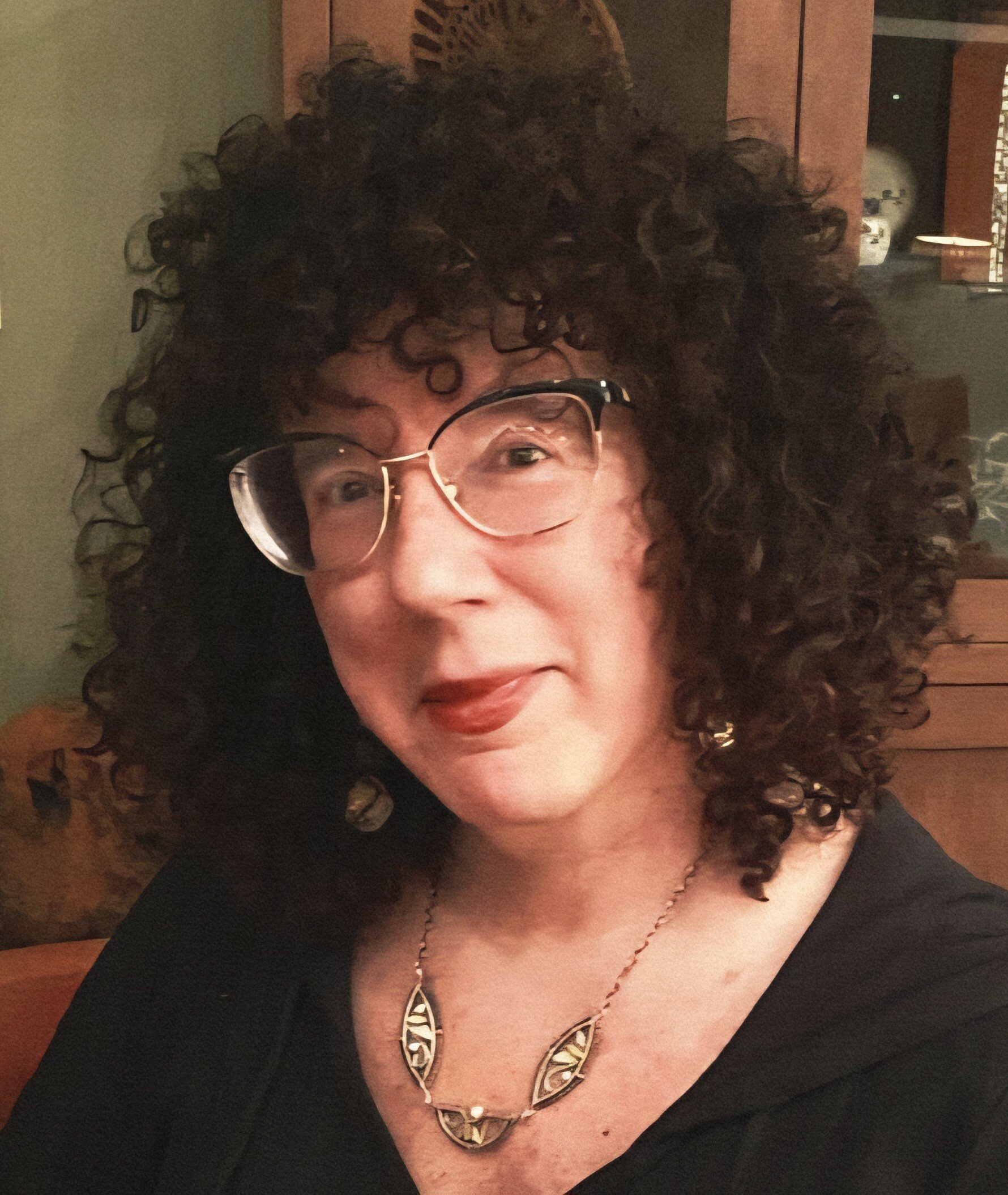 A woman with curly dark hair wearing glasses, a black top, and a gold necklace, smiling in an indoor setting.