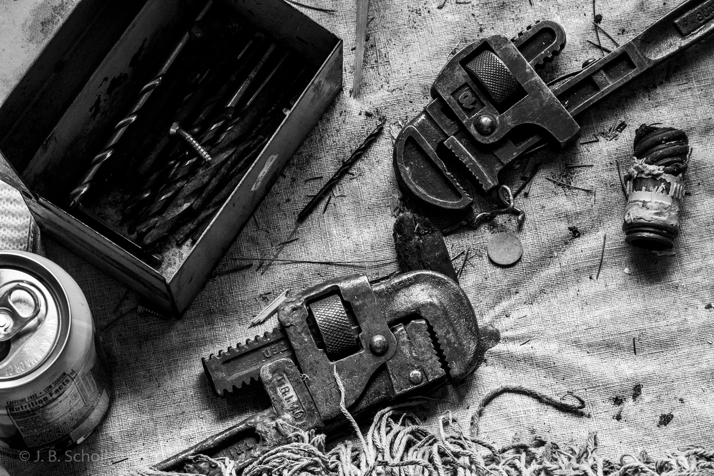 Black and white photo of assorted tools and objects on a textured surface, including a drill, a saw, a rope, and a can.
