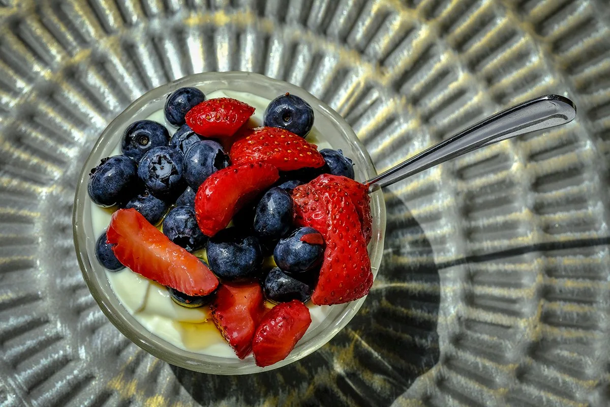 A glass bowl filled with strawberries and blueberries on top of a metallic surface, with a spoon inside the bowl.