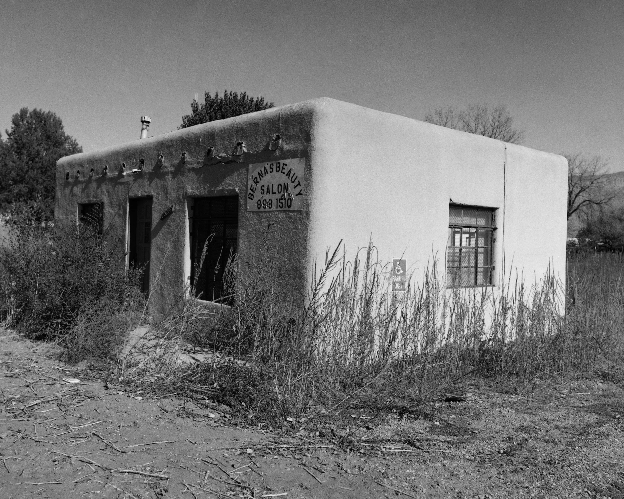 A small, adobe-style building with a sign for 'Bernas Beauty Salon' and a visible phone number, surrounded by overgrown grass and bushes, with a clear sky in the background.