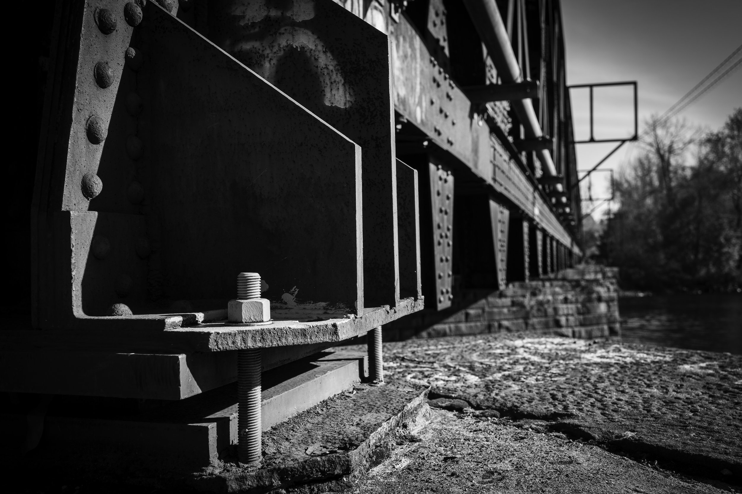 Close-up of a steel bridge's riveted metal support beams and bolts, with a blurred background of the bridge's structure, trees, and clear sky in black and white.