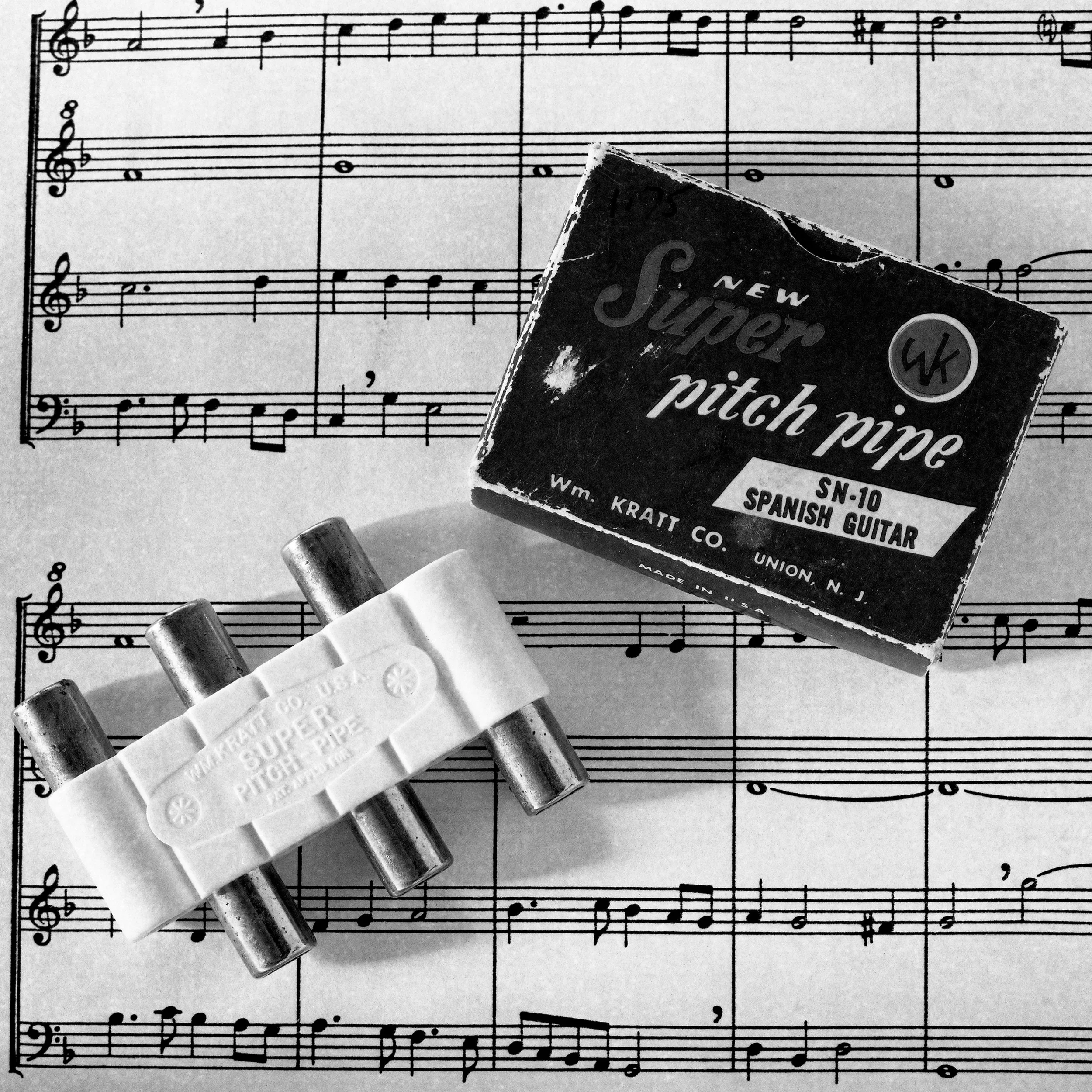 A sheet of music with musical notes, three guitar slides with metal barrels, and a small box of new super pitch pipe attached to one of the slides.