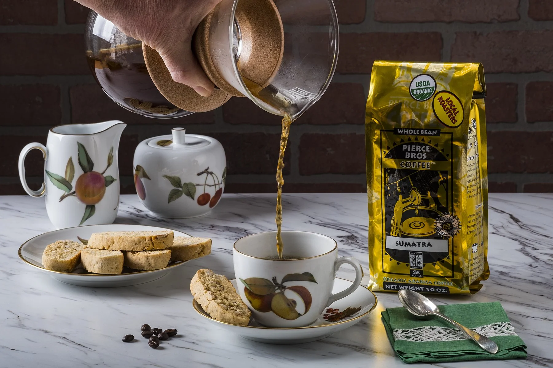 Pouring coffee into a cup from a glass coffee pot. There are cookies on a plate, a bag of Pierce Bros Coffee, a teapot, and a cup and saucer with a floral design on a marble table. Coffee beans and a spoon on a green napkin are also visible.