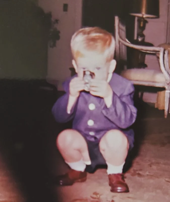 A young boy with light hair sitting on the floor, holding a camera or toy up to his eye, wearing a purple shirt with white buttons, shorts, white socks, and brown shoes, in a room with a wooden floor, a wooden chair, and a candle holder in the background.