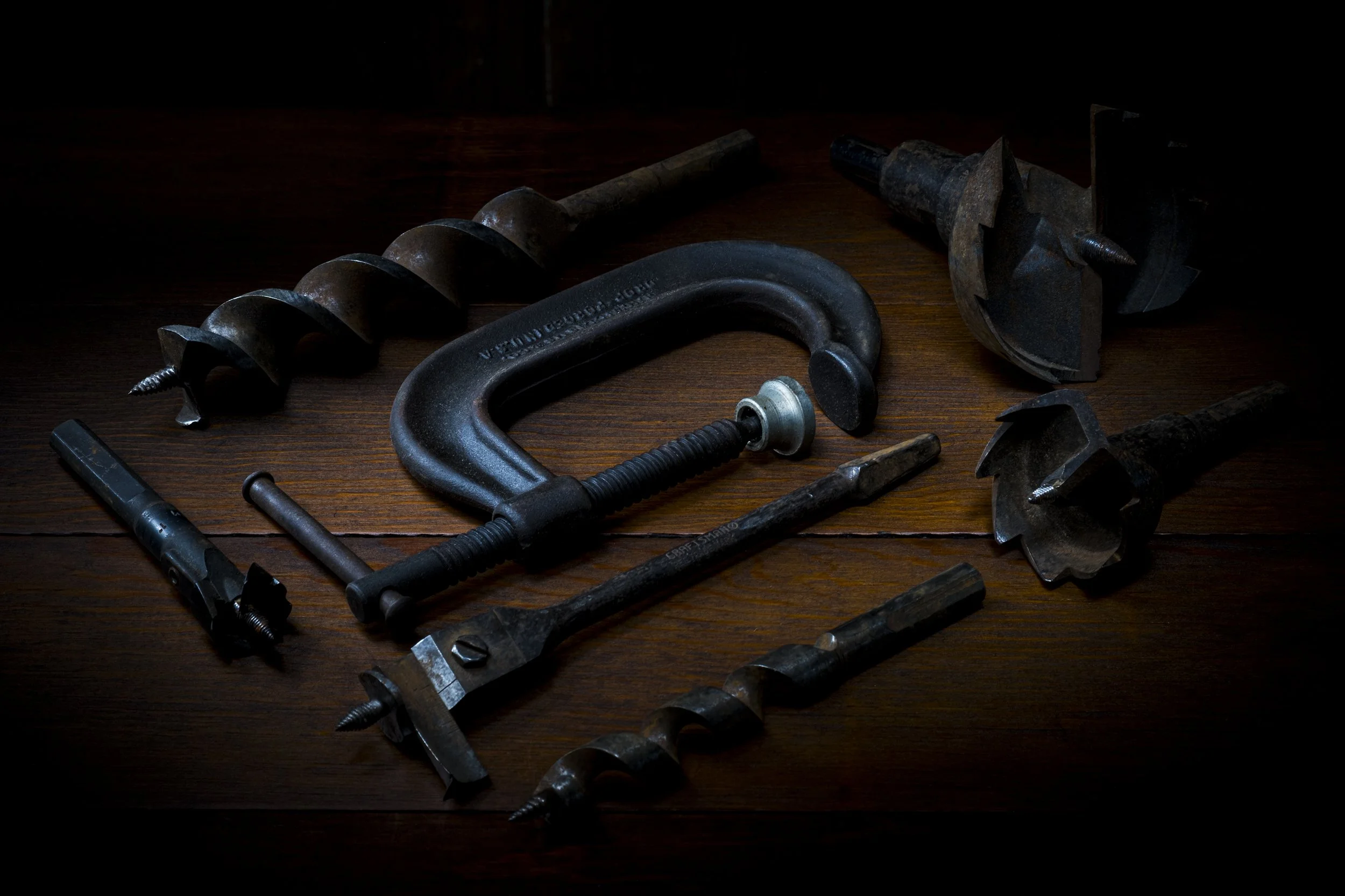 Various rusted metal tools including a corkscrew, a C-clamp, a pipe wrench, and screwdrivers are scattered on a dark wooden surface.
