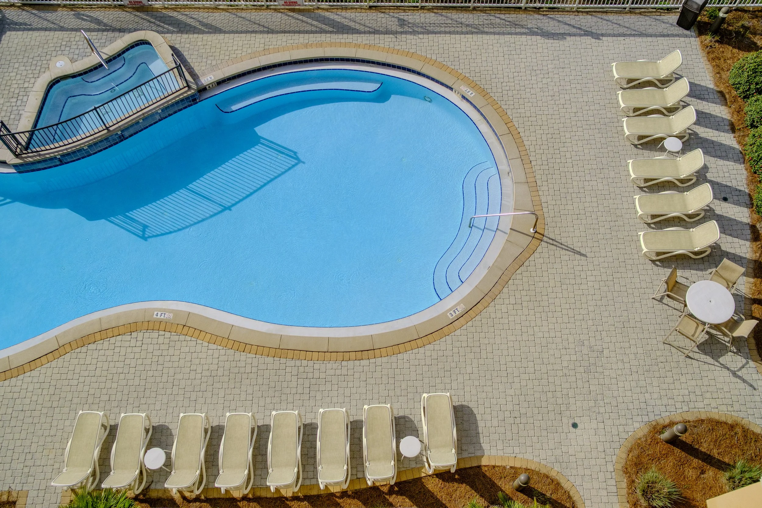 An aerial view of a swimming pool with a smaller hot tub connected via a narrow channel, surrounded by a paved pool deck with beige lounge chairs and small white tables.