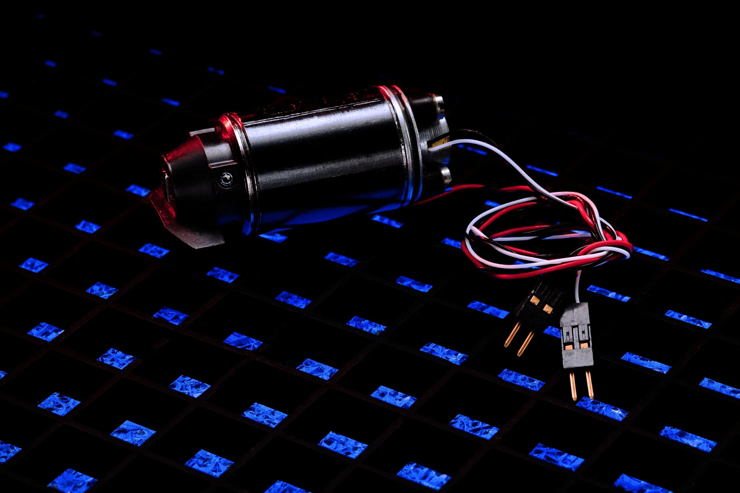 A small electric motor with red, white, and black wires connected, resting on a dark surface with a pattern of blue illuminated squares.