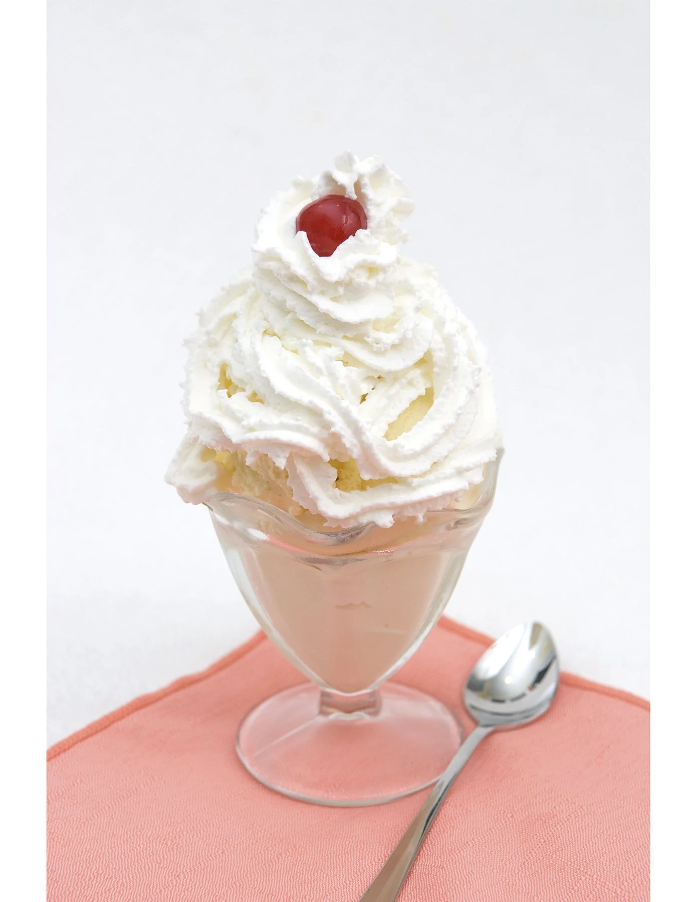 Icecream dessert in a glass cup with whipped cream topping and a cherry on top, served on a pink napkin with a silver spoon nearby, against a white background.