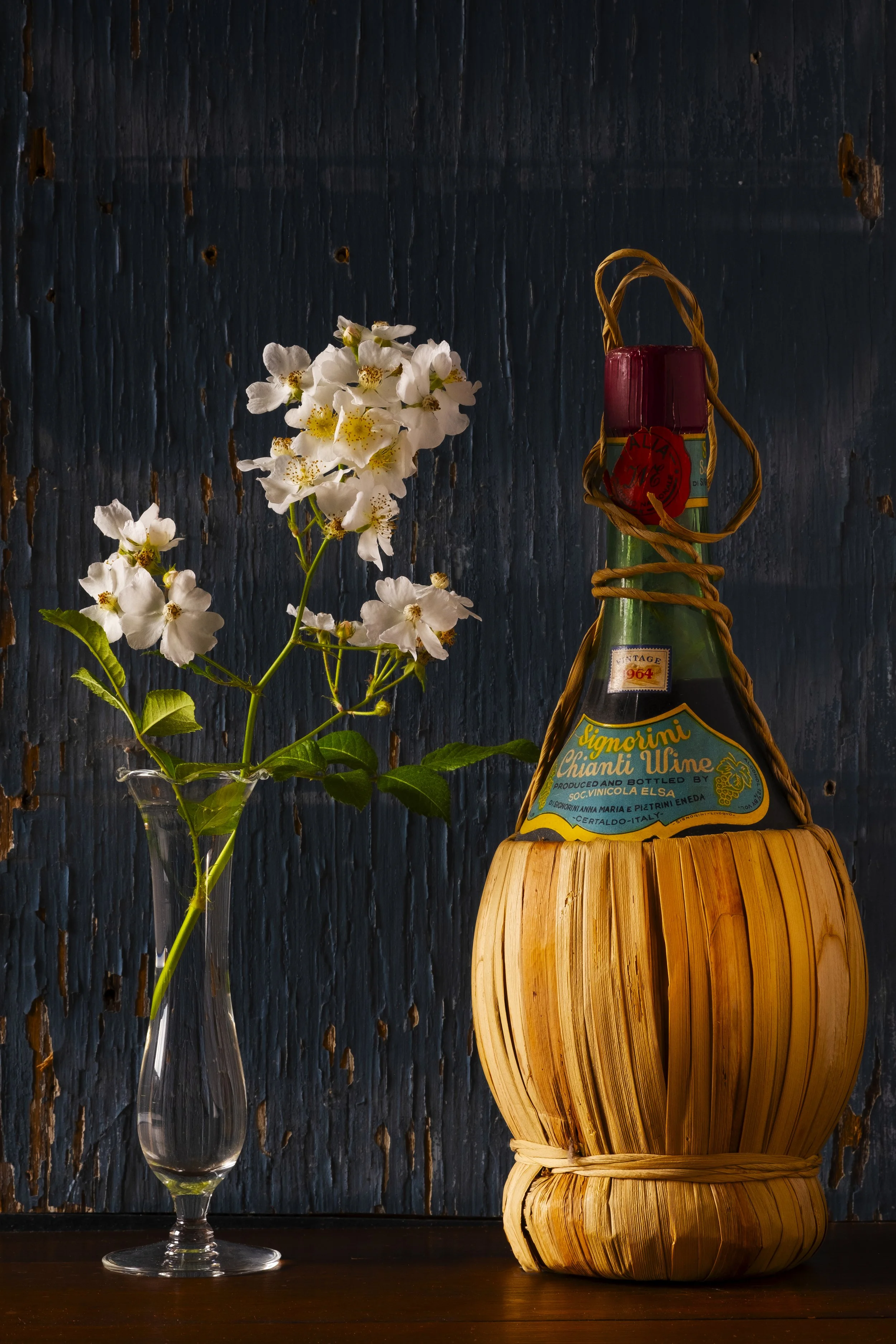 A clear glass vase with white flowers next to a bottle of Chianti wine wrapped in a bamboo covered container, set against a dark wooden background.