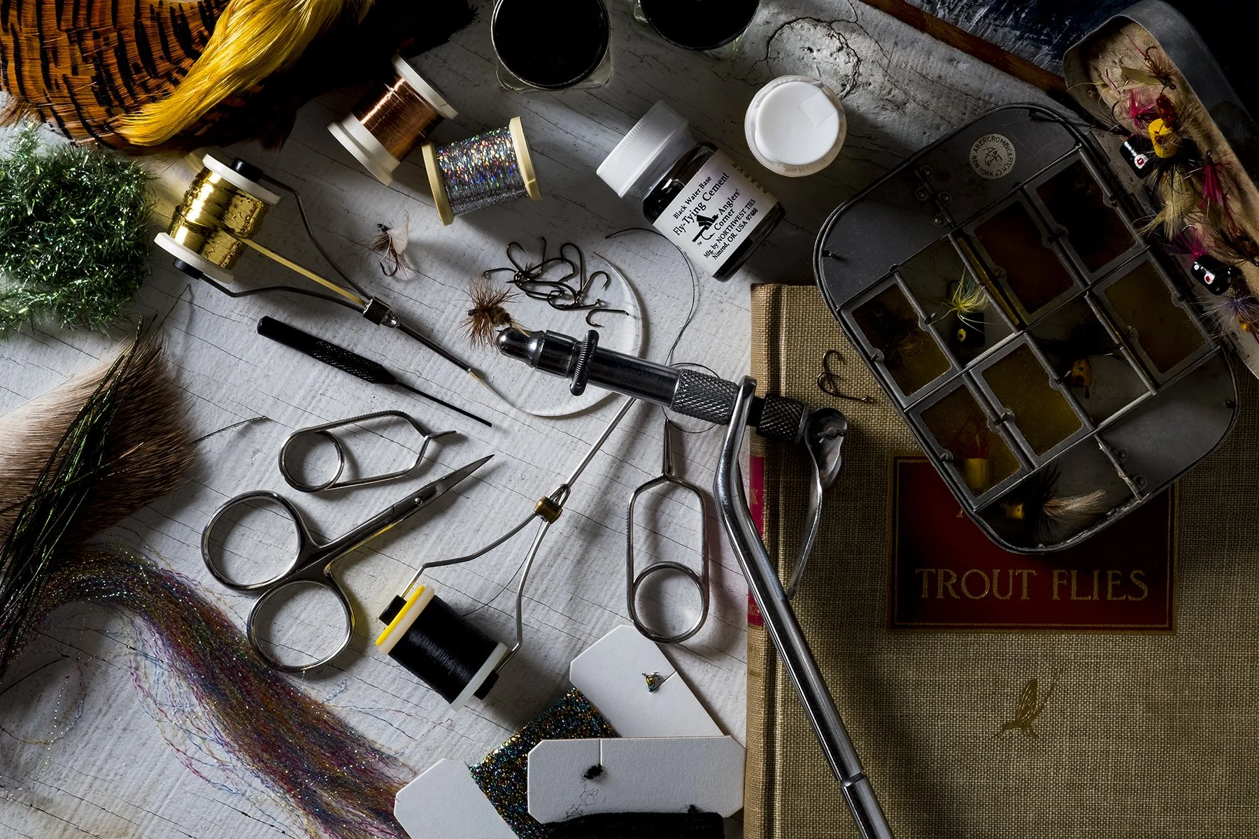 A collection of fly tying tools and materials, including scissors, thread spools, feathers, beads, and a fly tying box, arranged on a light-colored wooden surface.