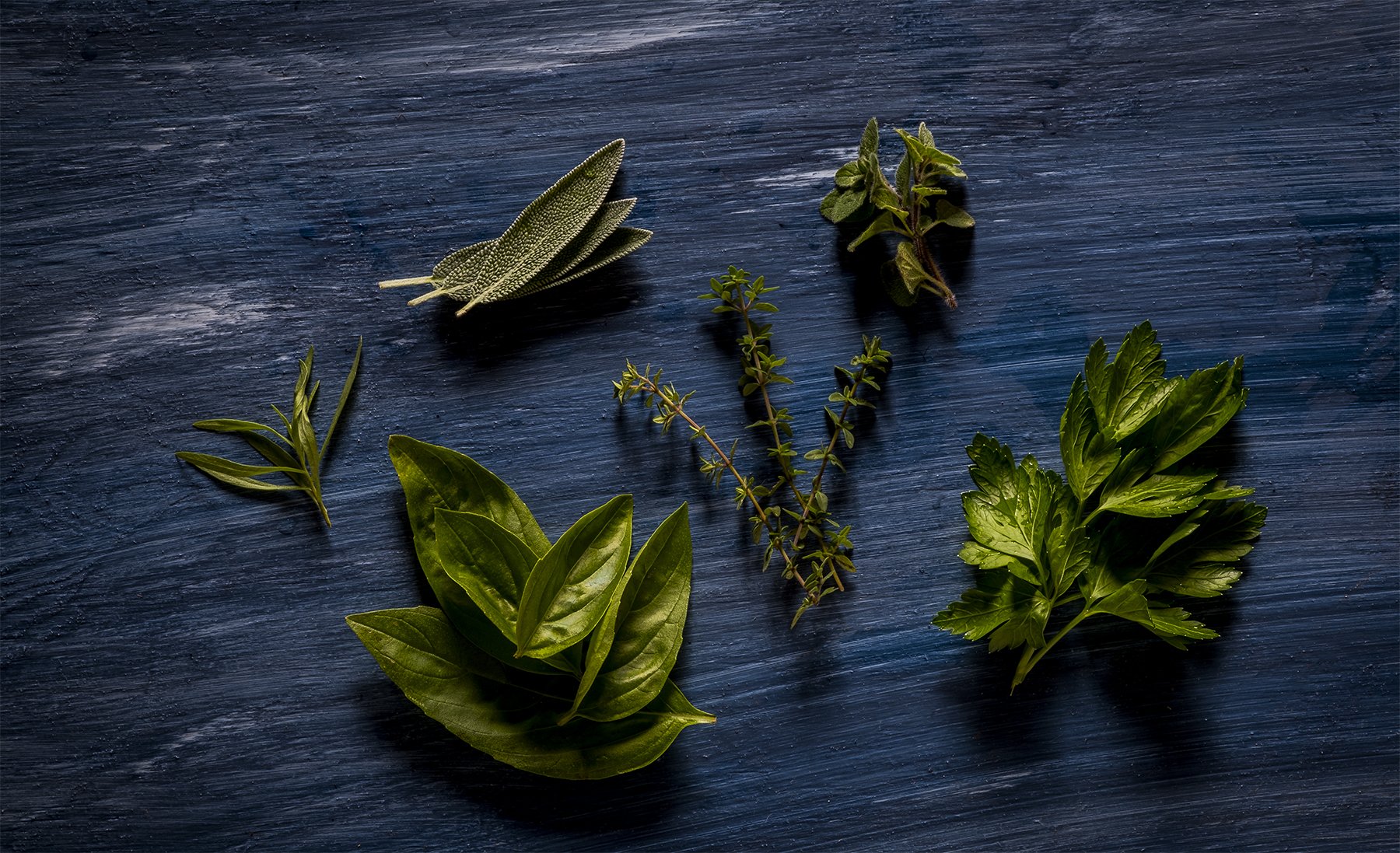 Various fresh green herbs and leaves arranged on a dark wooden surface, including sage, thyme, parsley, basil, and tarragon.