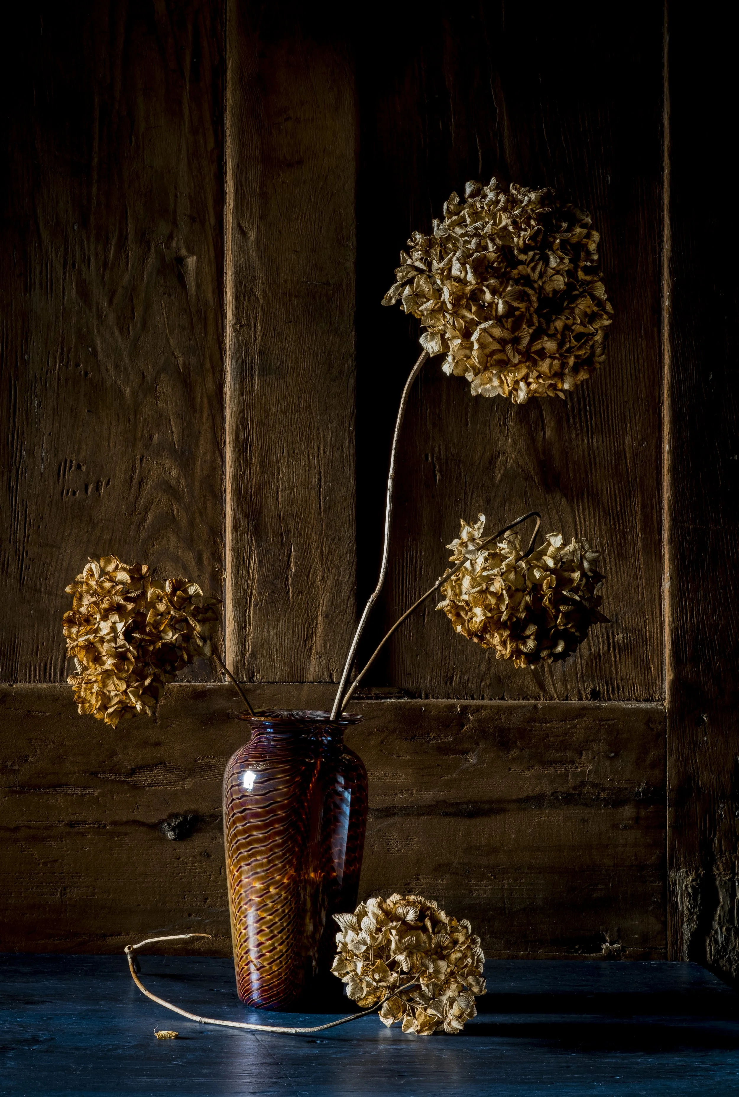 A tall, multicolored glass vase with a copper, orange, and purple swirl pattern, holding dried hydrangea flowers with beige petals, set against a dark wooden background on a black surface.