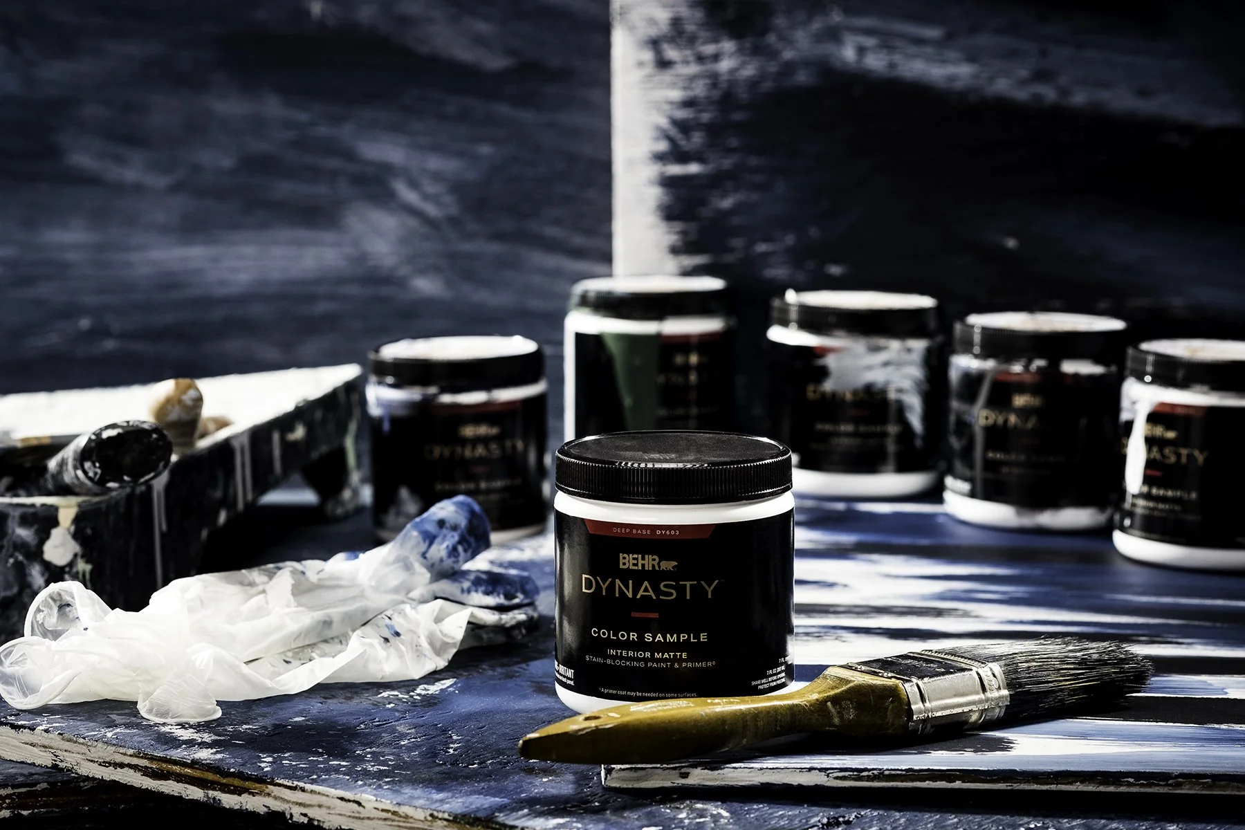 Multiple black containers of BEHR Dynasty interior matte stain-blocking paint and primer on a work surface, with artist brushes, a paint roller, and crumpled gloves, set against a painted blue wall background.