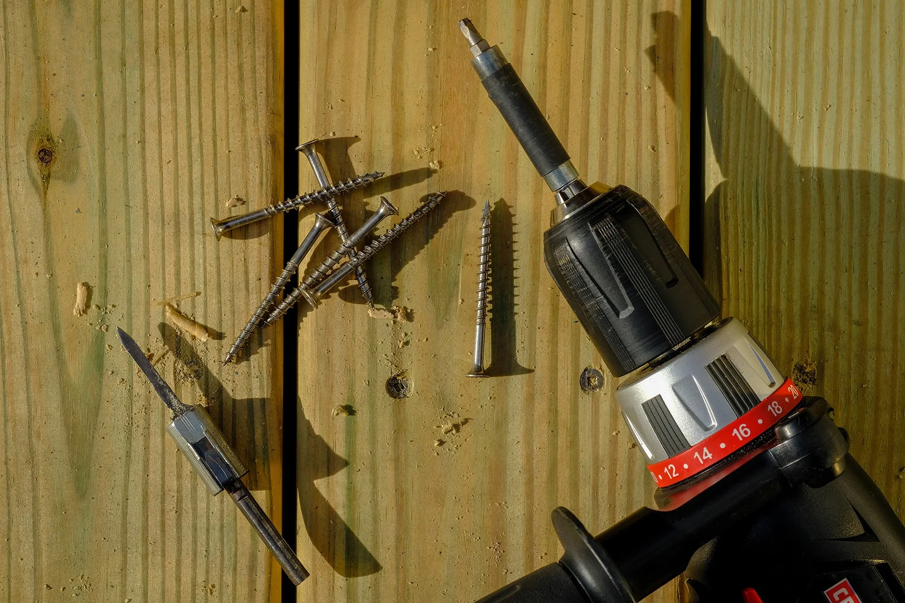 A cordless power drill, used for screwing, lying on a wooden surface along with several screws, a screwdriver, and a small piece of wood.