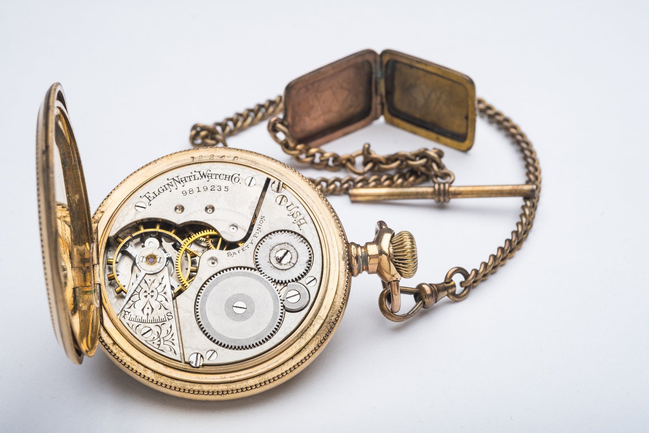A vintage gold pocket watch with the cover open, revealing the intricate mechanical inner workings, along with a matching chain and a small metal tool, all placed on a white surface.