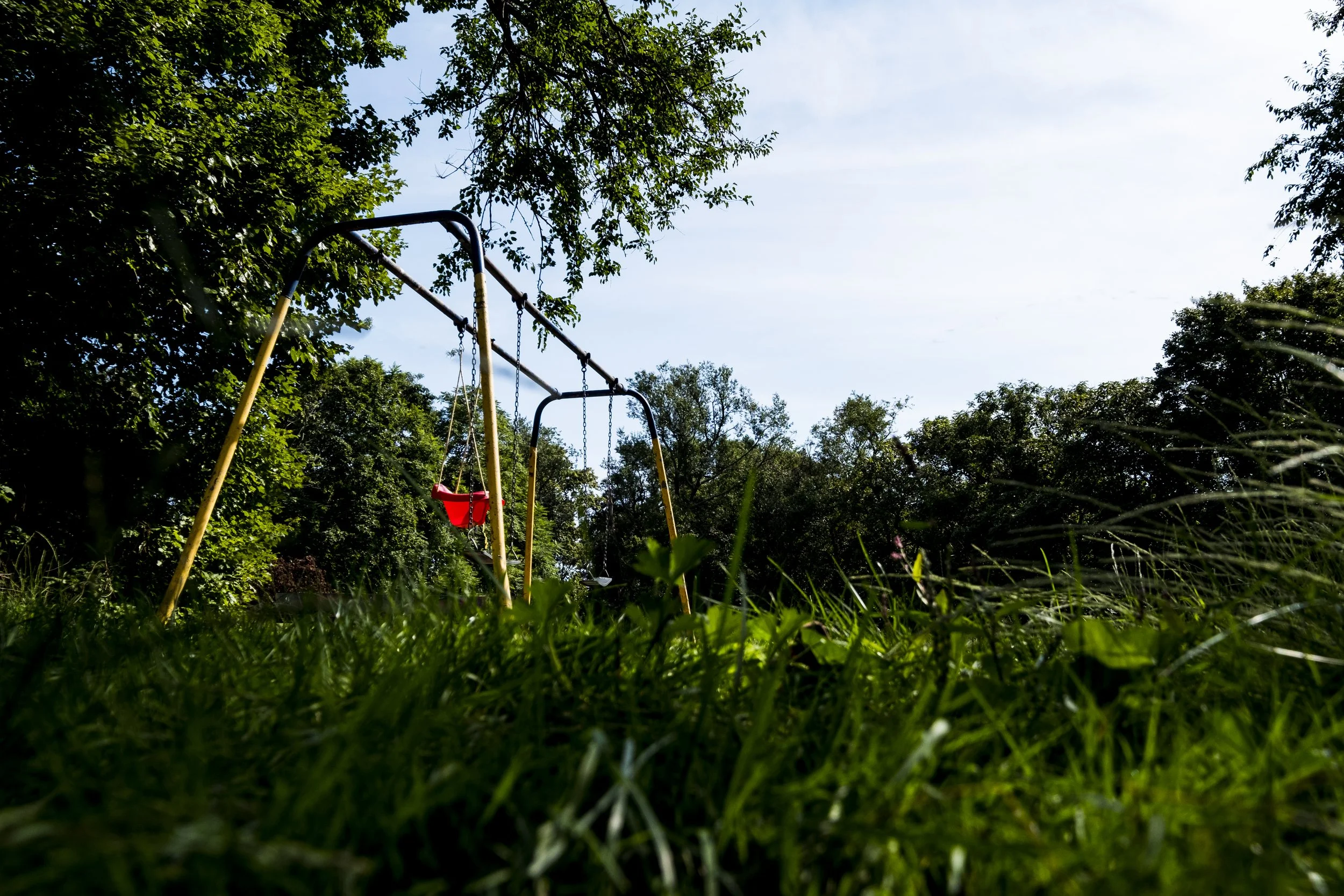 A playground swing set with the two swings, one red and one silver, surrounded by green trees and grass under a blue sky.