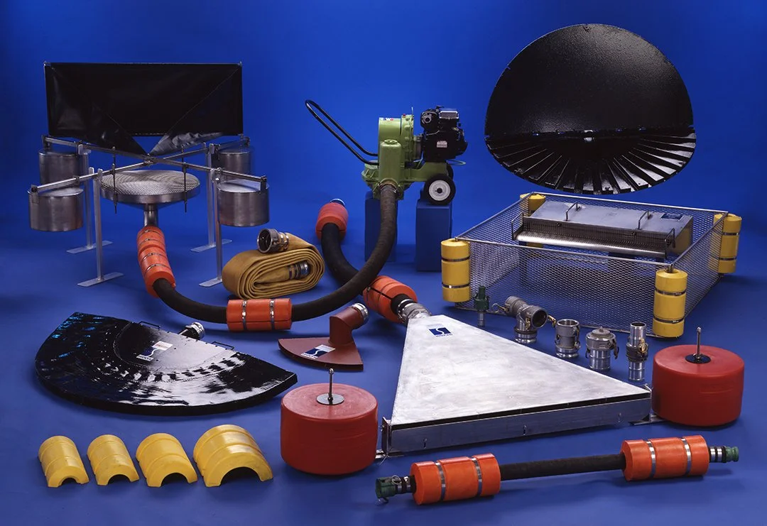 Various oil slick removal equipment and tools laid out on a blue surface, including hoses, nozzles, a large metal scoop, a yellow protective barrier, a red foam buoy, and a black fan shaped oil vacuum head.