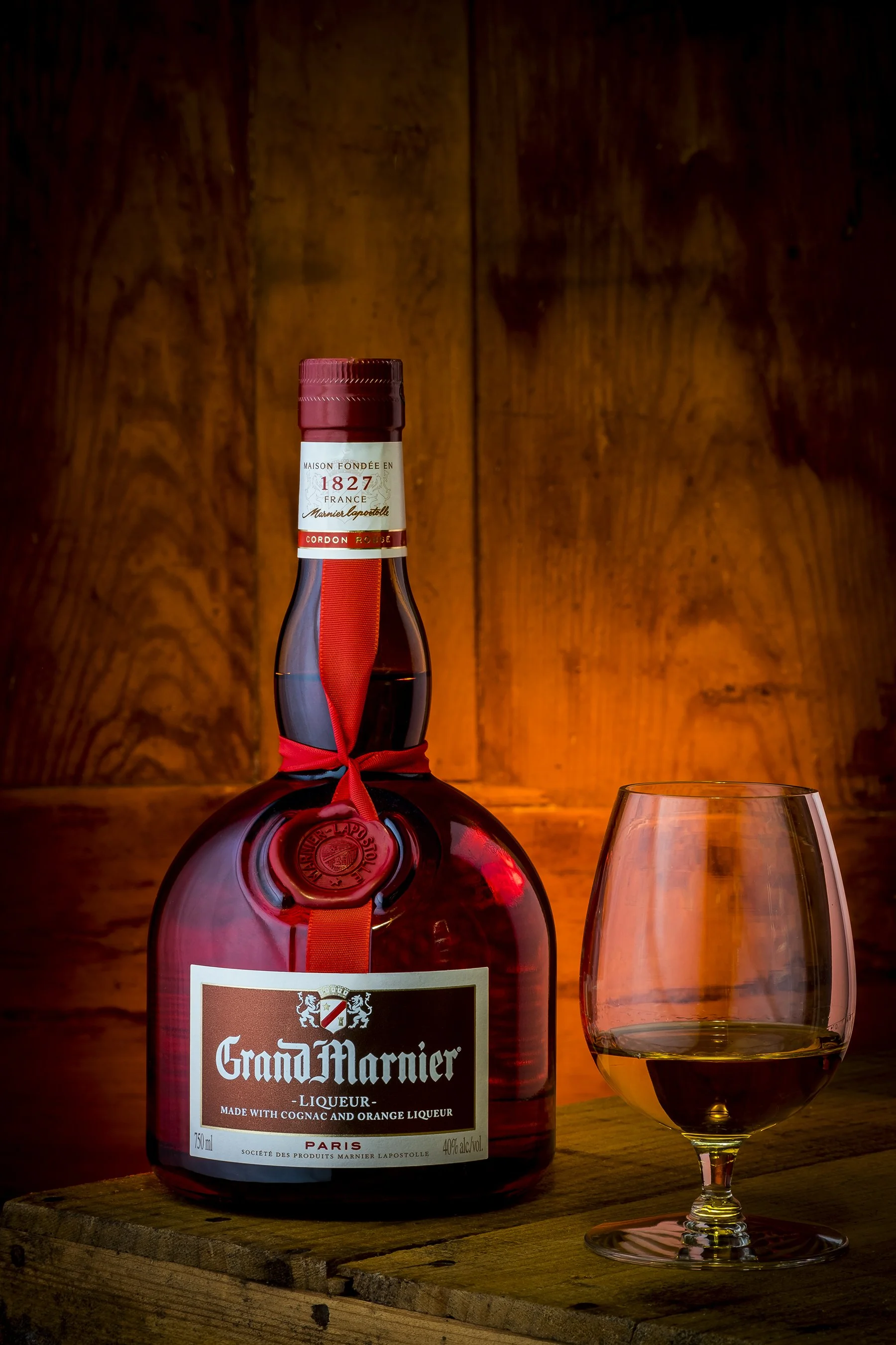 A bottle of Grand Marnier liqueur with a glass of dark liquor on a wooden table, against a wooden wall background.
