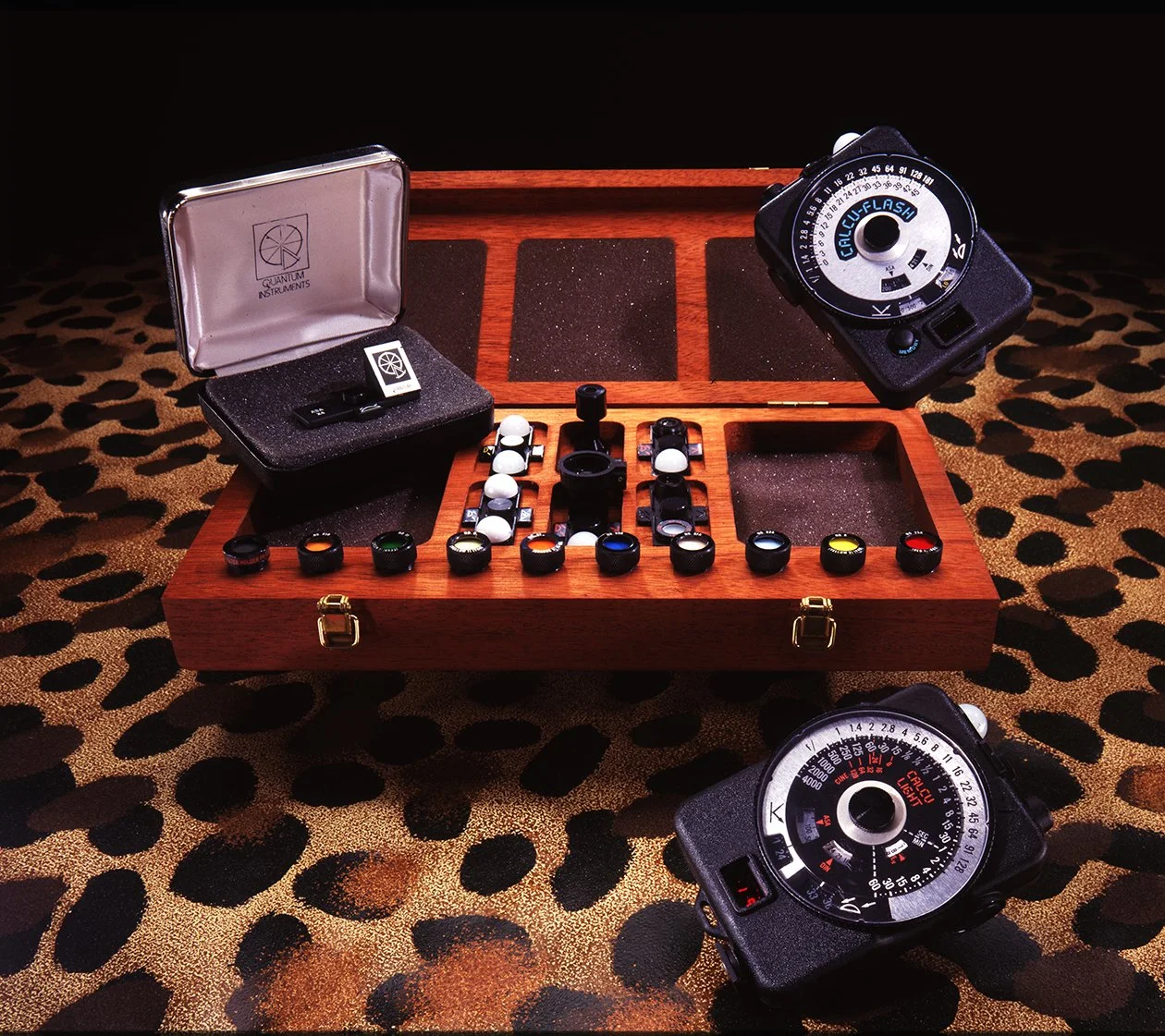 A collection of photographic filters and related equipment for early digital lightmeter displayed on a wooden case, with a leopard print fabric surface underneath.