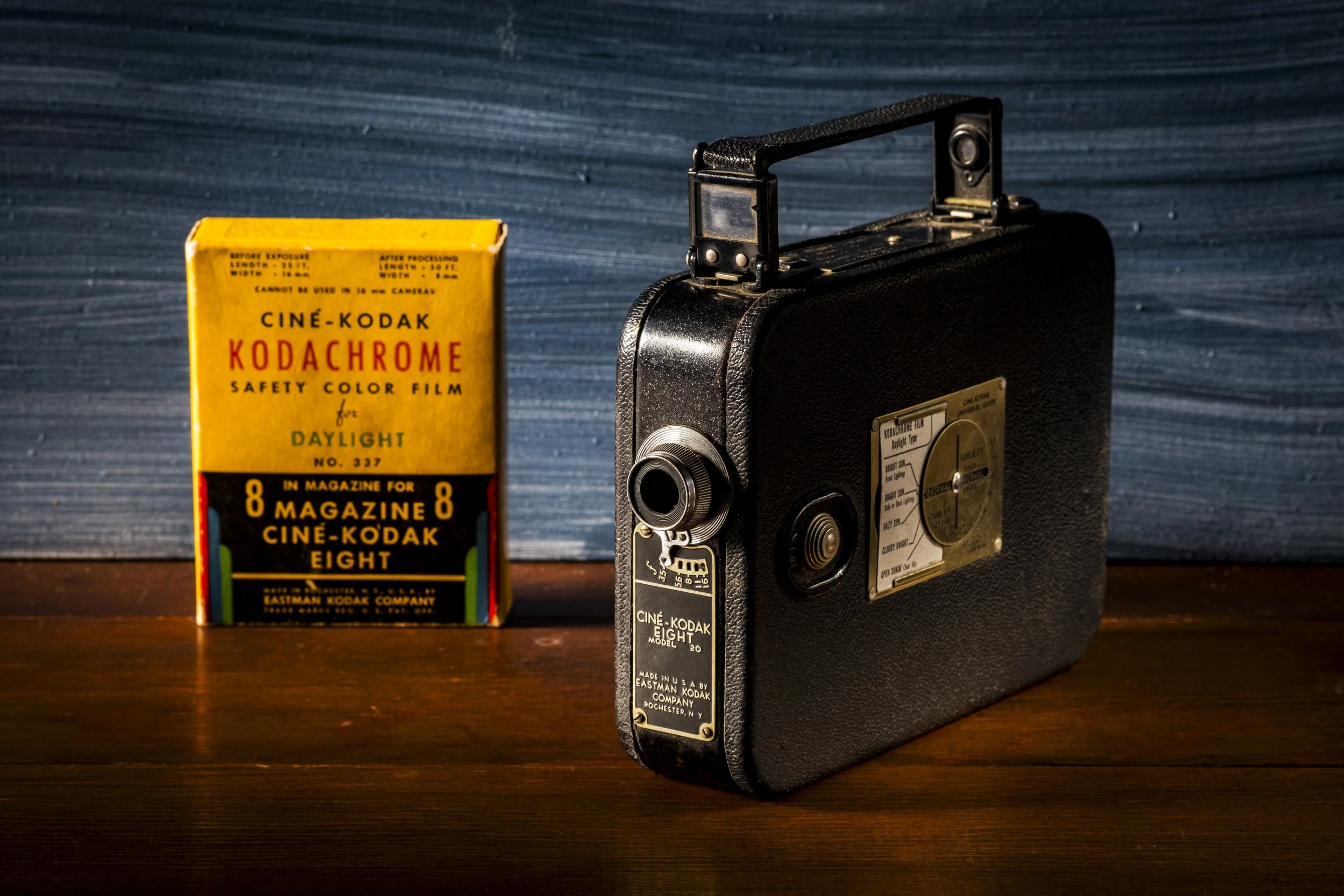 A vintage black film camera with a textured leather exterior and metal controls, positioned on a wooden surface. To the left, a yellow box of Kodak Kodachrome color film for eight magazine prints stands upright against a blue wooden background.
