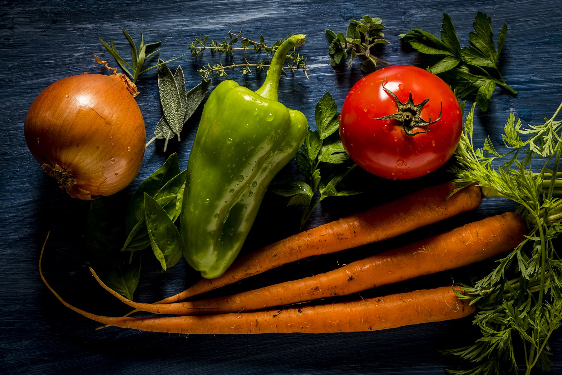 Fresh vegetables including an onion, a green bell pepper, a tomato, two carrots, and various herbs on a dark wooden surface.