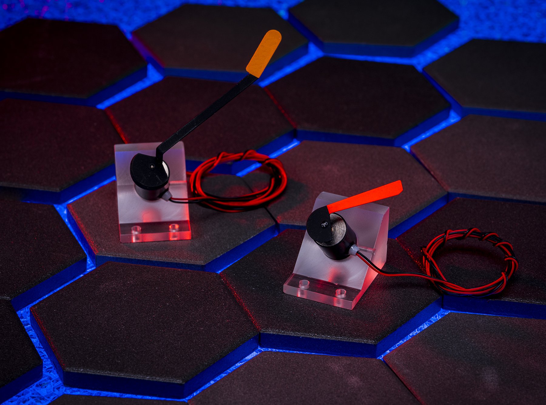 Two solenoid sensors with orange and red tips on a black hexagonal tiled surface with blue accents, connected by red and black wires.