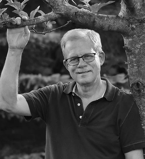 A middle-aged man with glasses and light-colored hair smiling, standing outdoors near a tree, holding a branch with one hand.