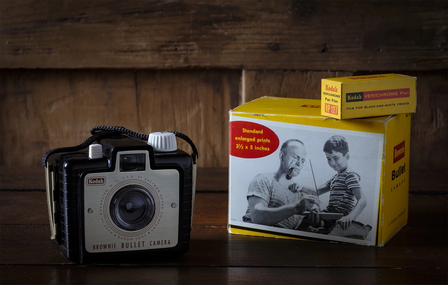 A vintage Kodak Brownie Bullet camera and a box of Kodak Verichrome Pan film, placed on a wooden surface against a wooden wall background.