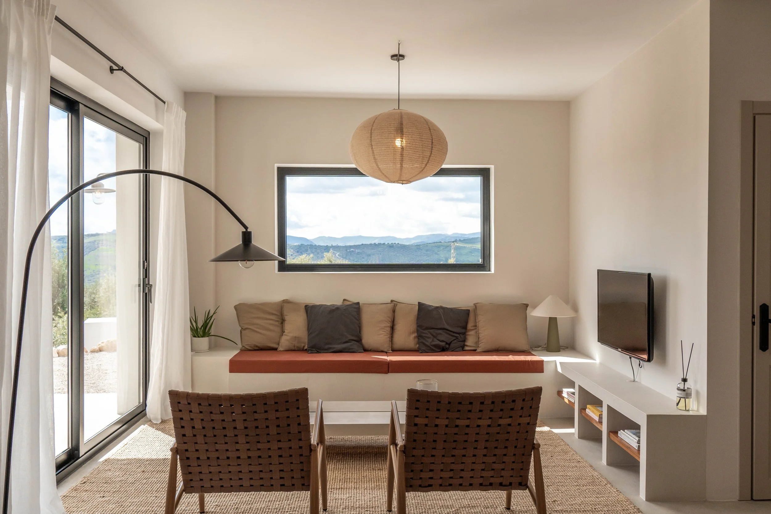 Interieur van een woonkamer met een loungebank, twee stoelen, een televisie, een schuifpui grenzend aan de veranda en een groot raam met uitzicht op een landschap met bergen.