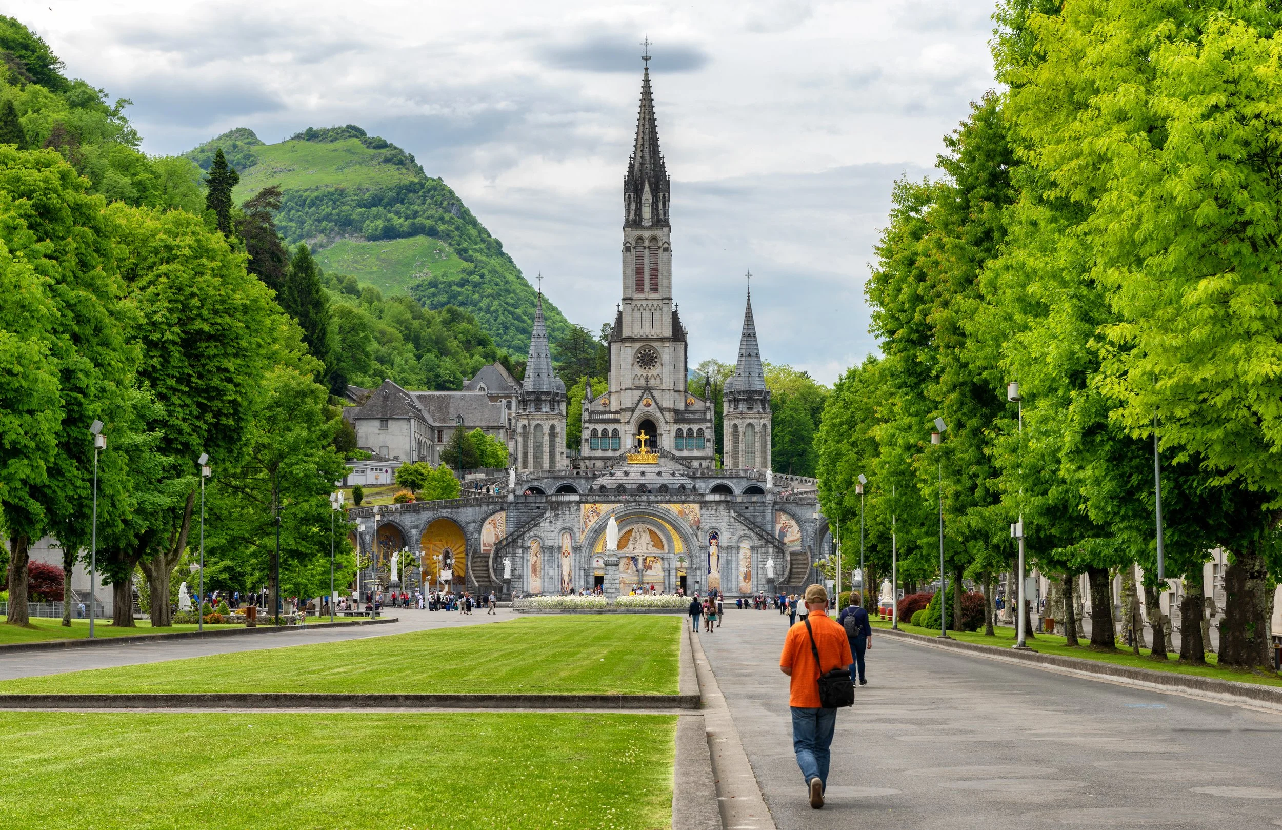 A large church with a tall steeple surrounded by lush green trees, hills in the background, and people walking on a paved pathway in the foreground.