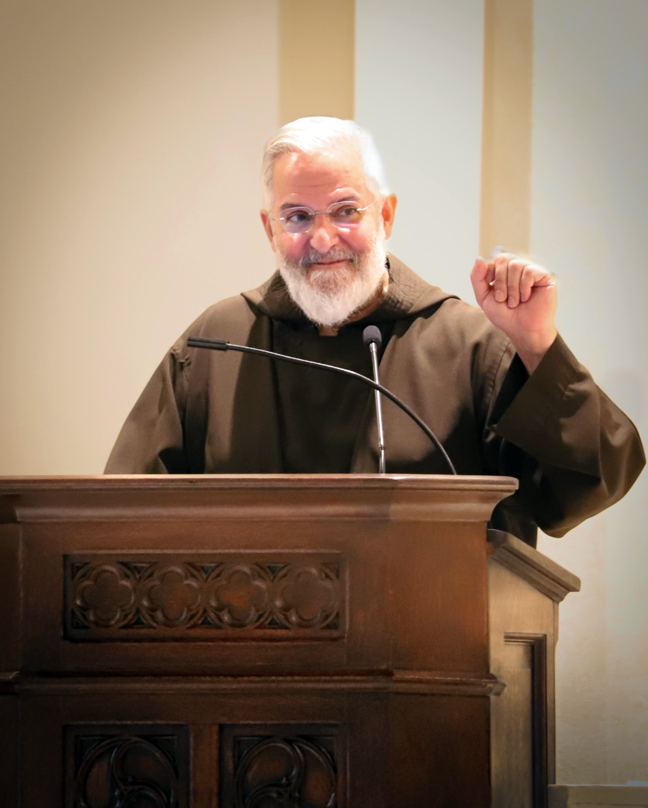 An elderly man with white hair and a beard, wearing glasses and a brown robe, standing at a wooden lectern with a microphone, raising his right fist in a gesture of triumph or encouragement, inside a church or similar setting.