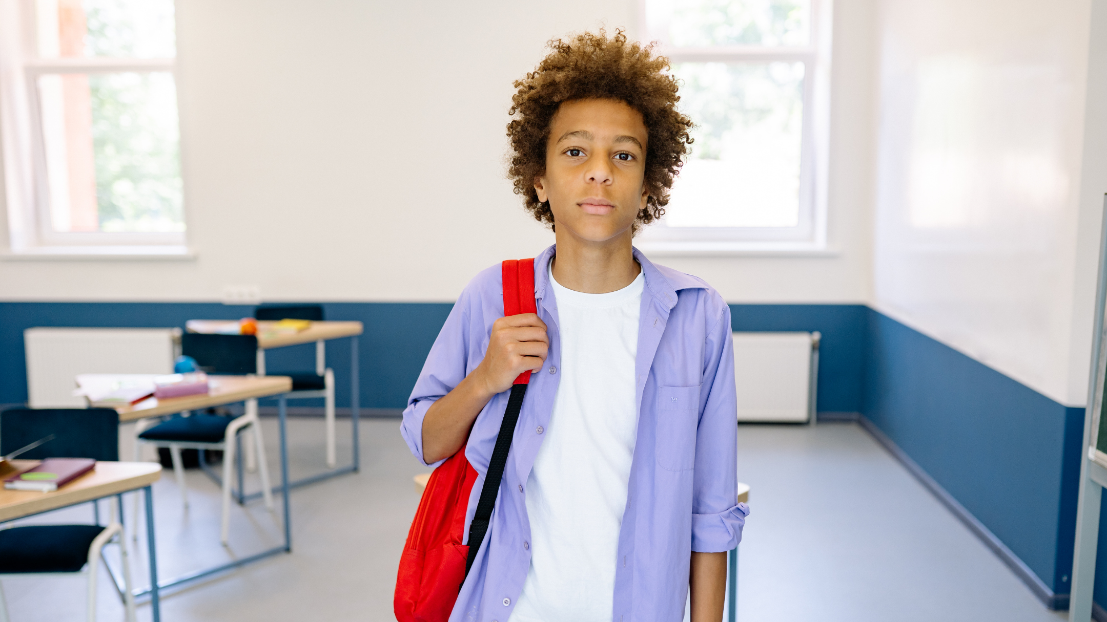 A student with curly hair and a red backpack standing in a classroom with desks and large windows.