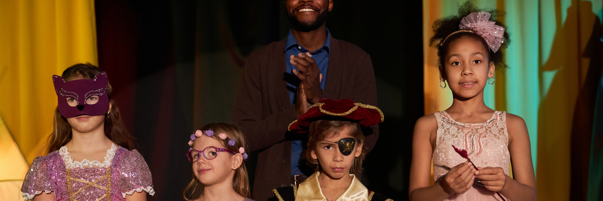 Children dressed in costumes on stage, with a man clapping in the background.