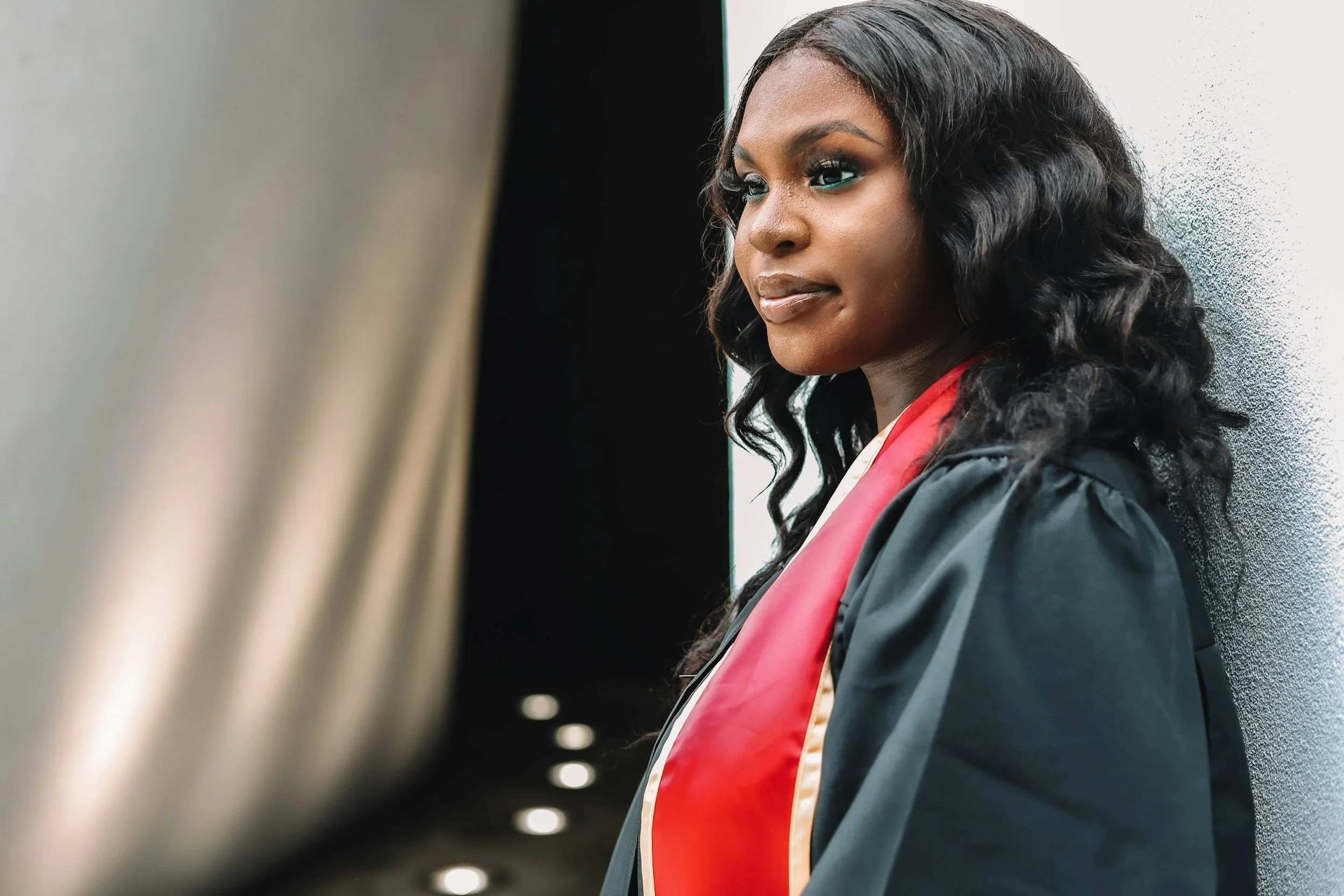 A woman in a black graduation gown and red sash standing against a grey wall, looking off to the side.