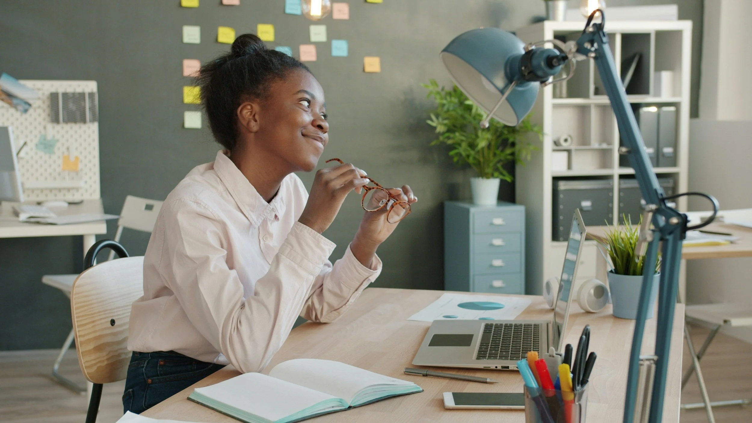 A young woman with dark hair tied back, wearing a light pink shirt, sitting at a desk in a modern office. She is smiling and holding her eyeglasses in red and tortoiseshell, with an open notebook, a laptop, and a tablet on the desk. The background includes a dark wall with colorful sticky notes, a green plant, and storage shelves.