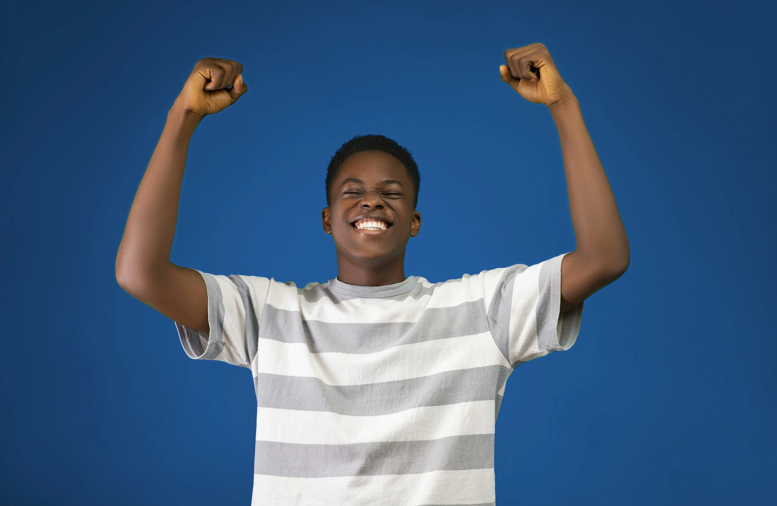 A young person with dark skin, short curly hair, smiling broadly with eyes closed, raising both fists in a gesture of triumph or celebration against a solid blue background, wearing a white and gray striped T-shirt.