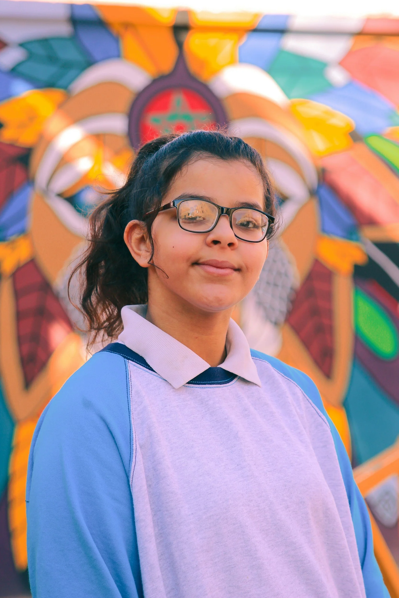 A young girl with glasses and a light purple and blue shirt smiling in front of a colorful amusement park ride background.