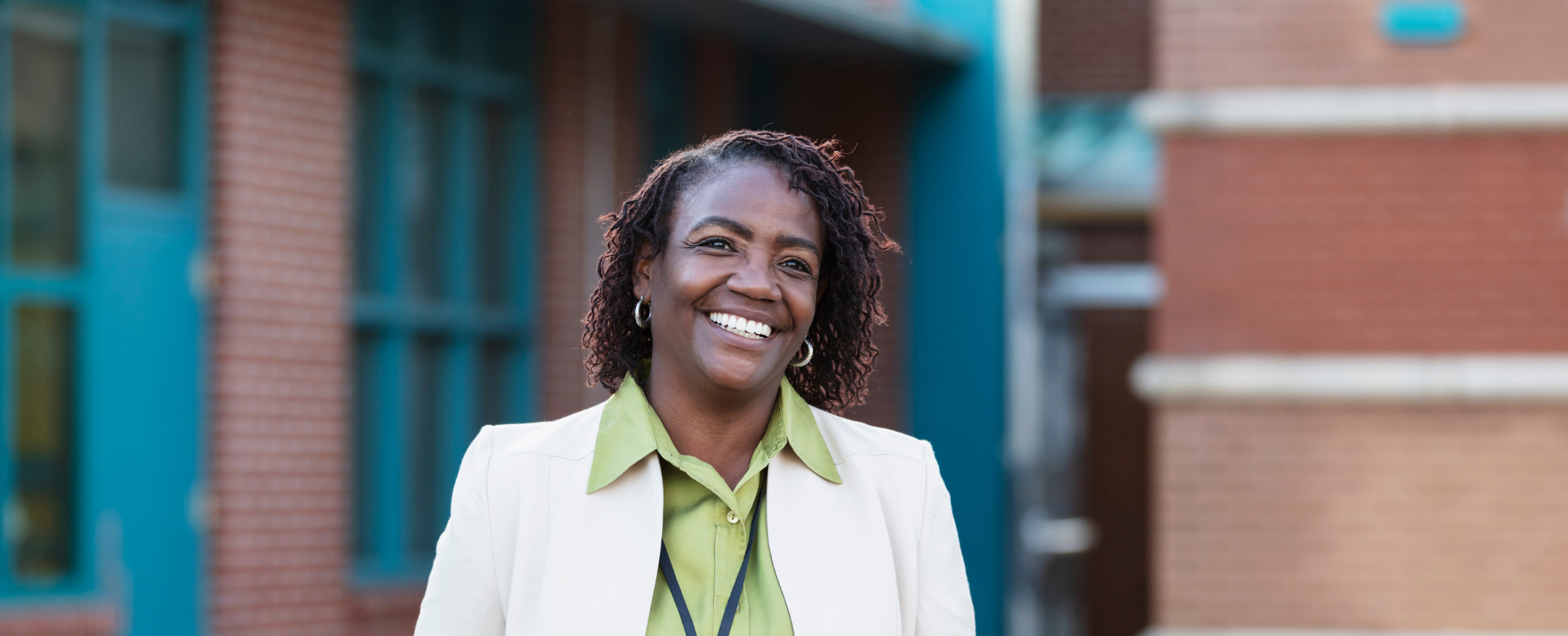 A smiling woman with dark curly hair wearing a white blazer and lime green shirt standing outdoors in front of a modern brick building.