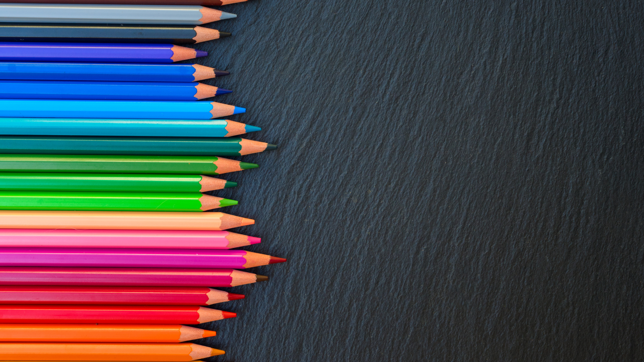 Line of colored pencils arranged on black textured surface, from pink to gray.