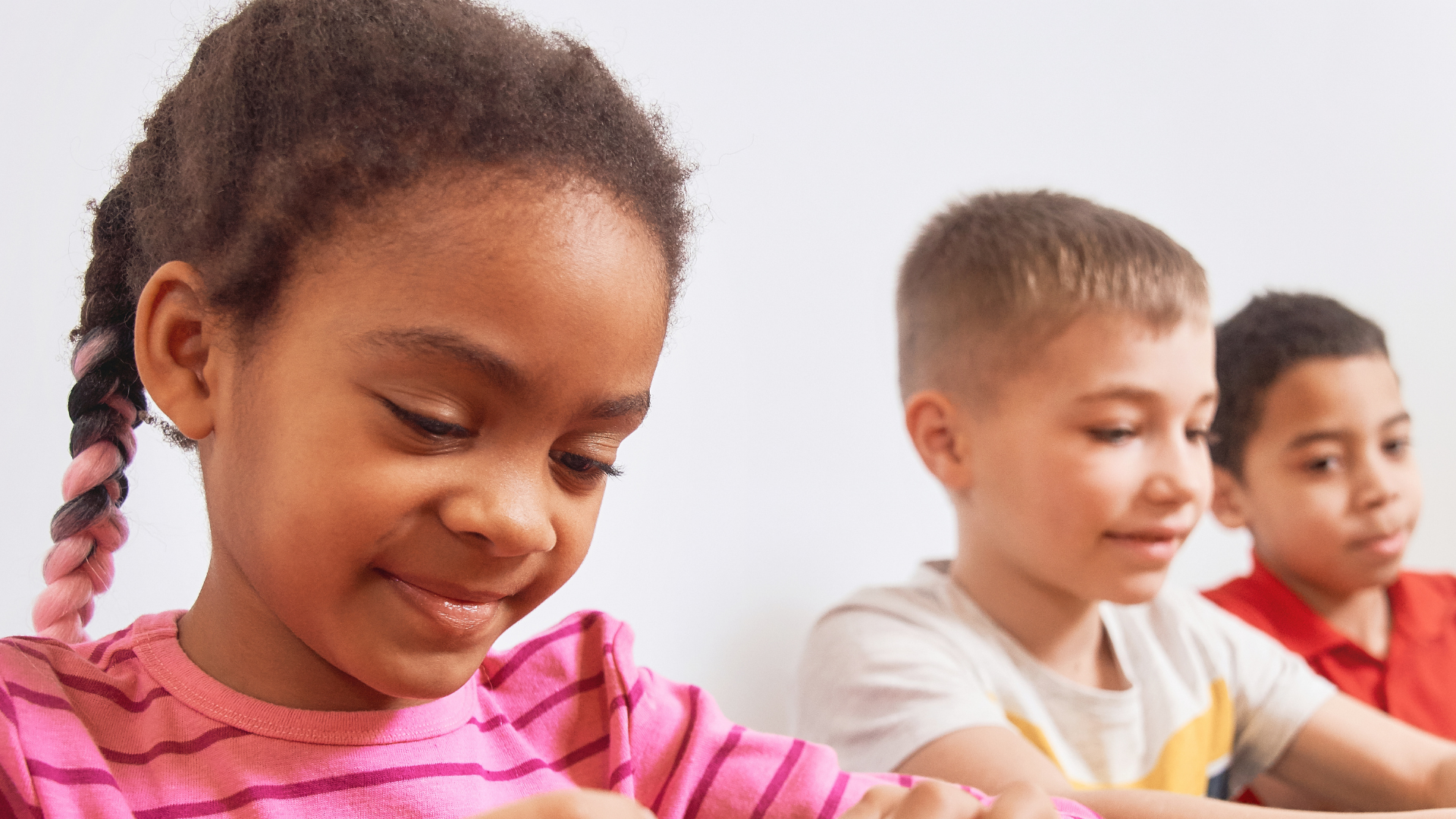 Three children sitting in a row looking downward, the girl in the foreground has brown skin, braided hair with pink extensions, and is wearing a pink and purple striped shirt. The two boys behind her have light brown skin, short hair, and are wearing beige and red shirts.