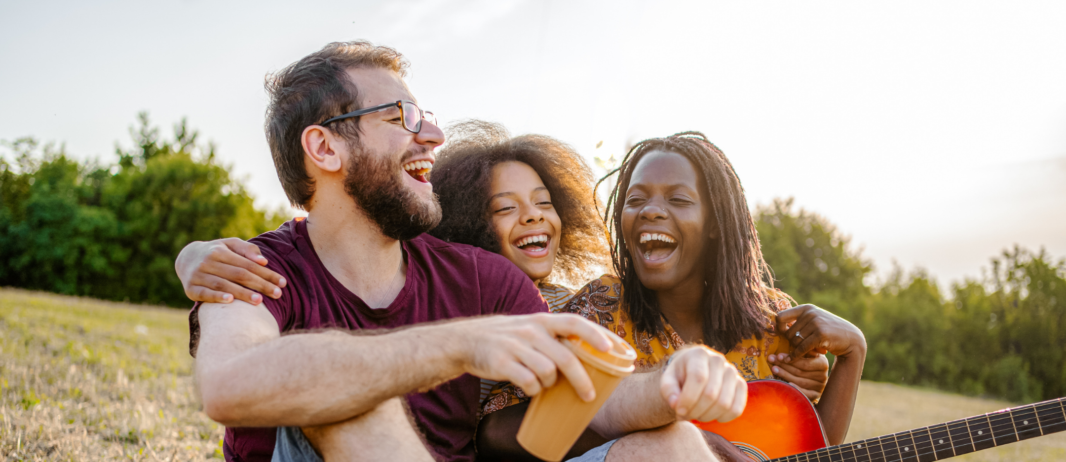 A group of three friends laughing and enjoying time together outdoors on a sunny day, with a guitar and a cup in hand.