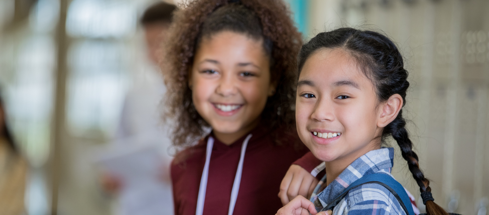 Two smiling young girls standing close together in a school hallway, one with curly hair in a maroon hoodie and the other with straight hair in a blue plaid shirt.