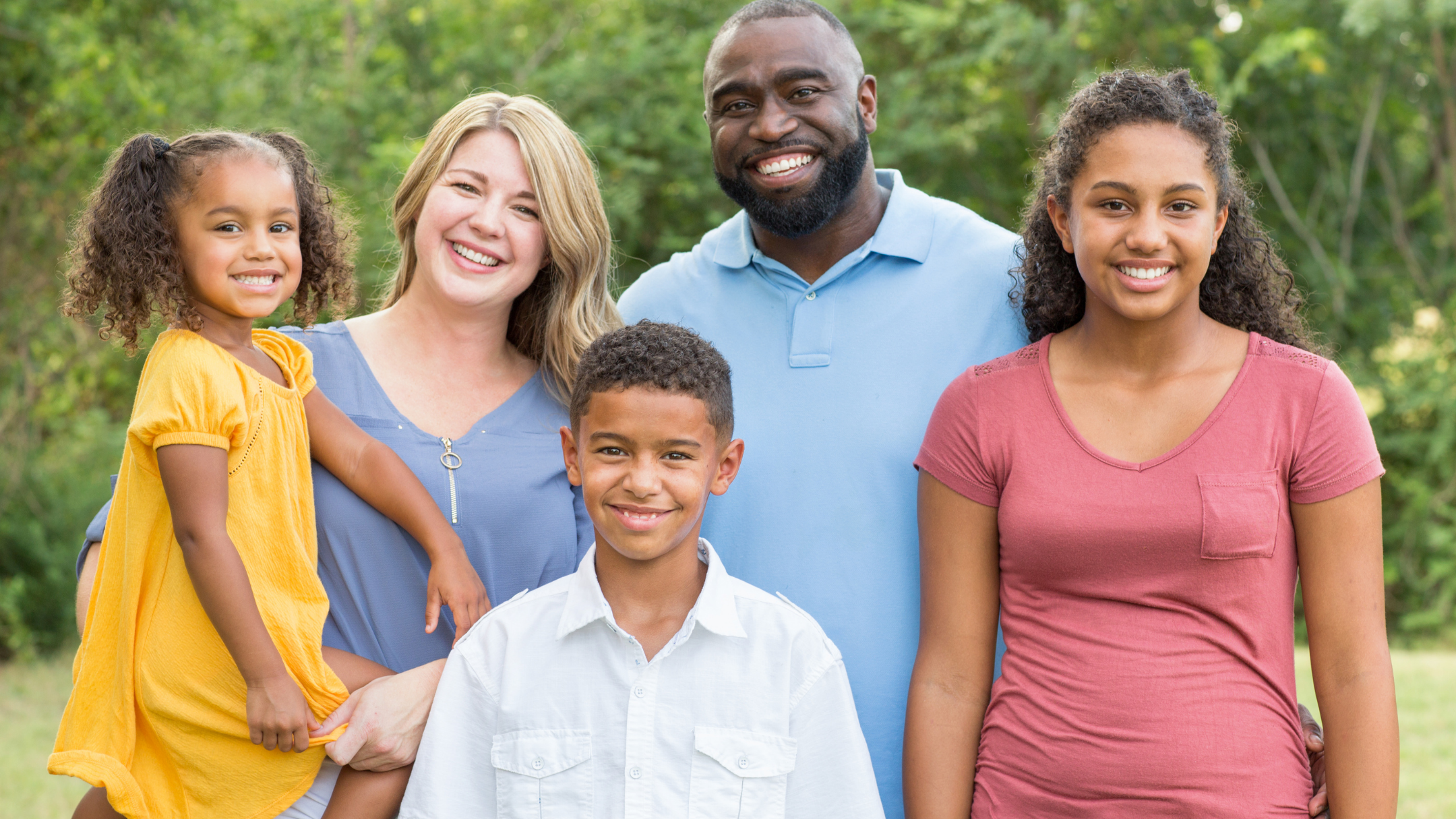 Group of five smiling people, including two young girls, two boys, and two adults, standing outdoors in a green park.